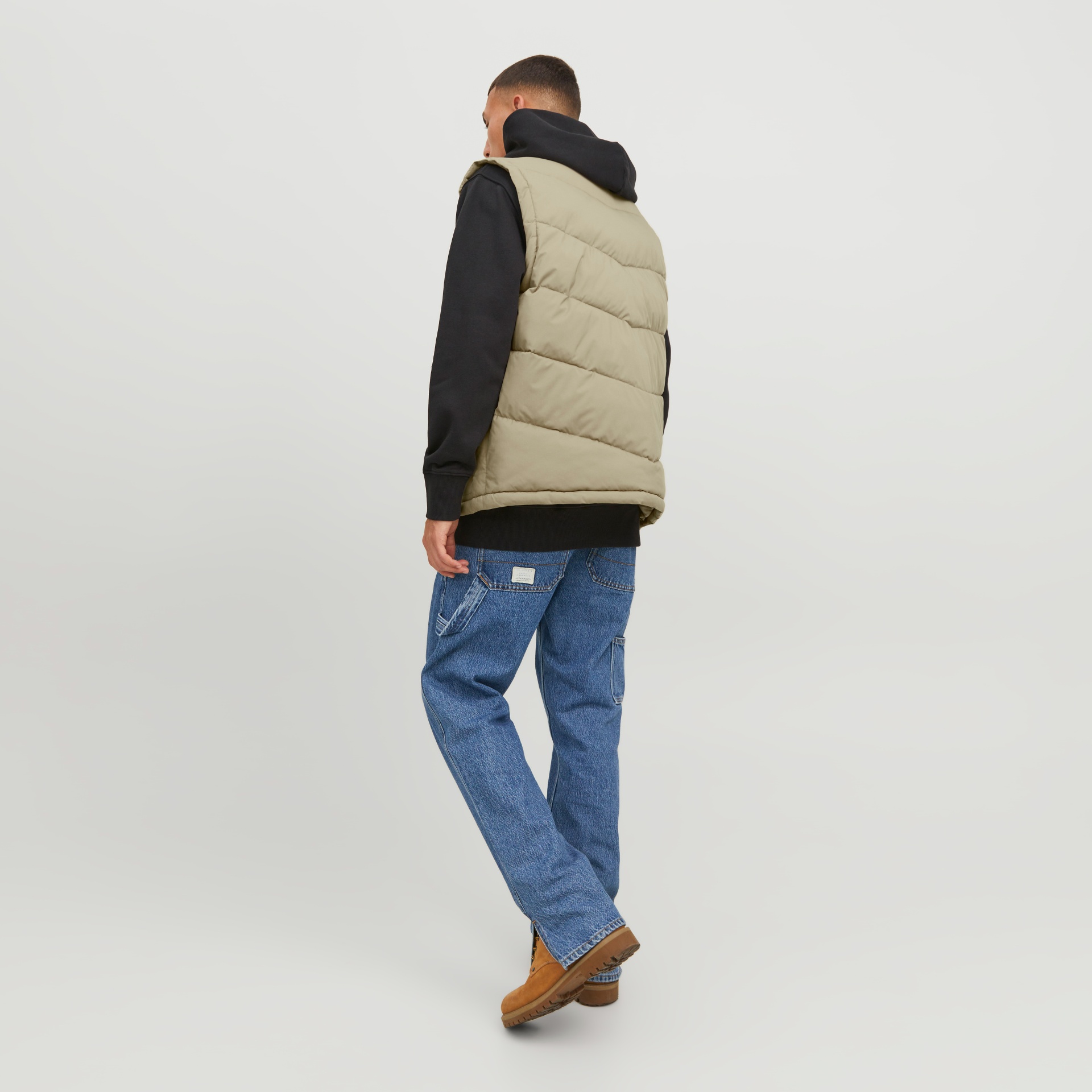 Homem de costas com colete bege, sweat preto e jeans azuis