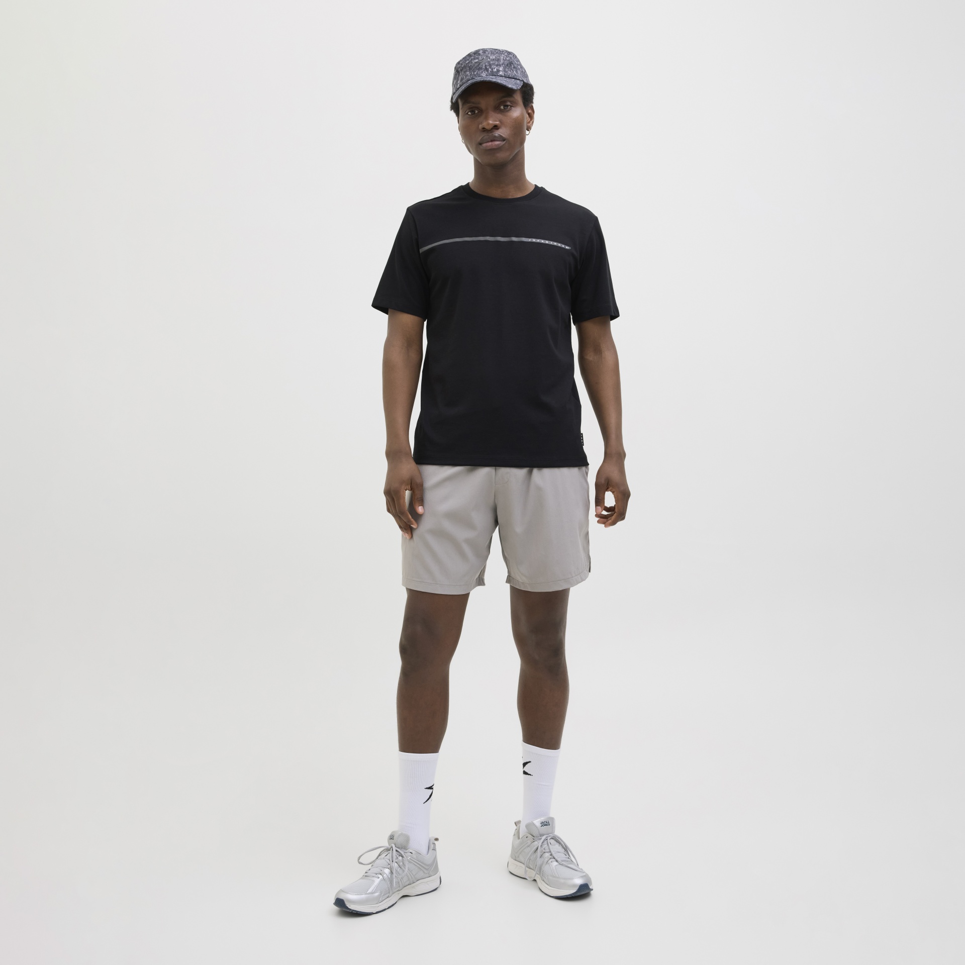 Homem vestido com t-shirt preta, calções cinza claros, boné cinzento escuro, meias brancas e ténis cinzentos