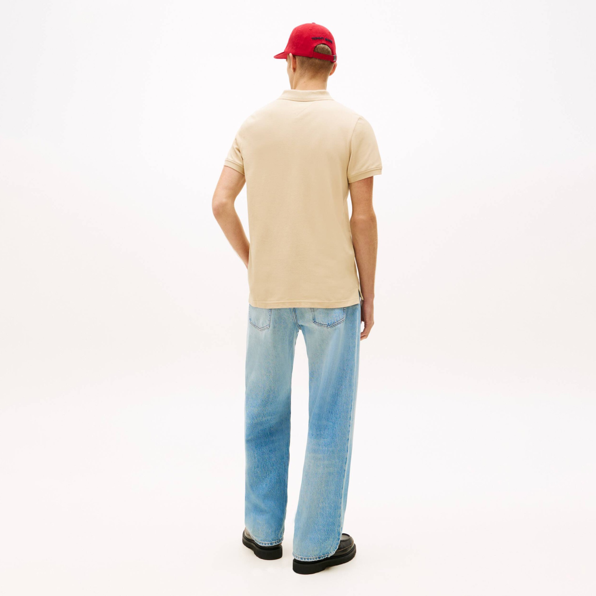 Homem de costas com t-shirt polo bege, calças de ganga azuis claras e boné vermelho Tommy Jeans