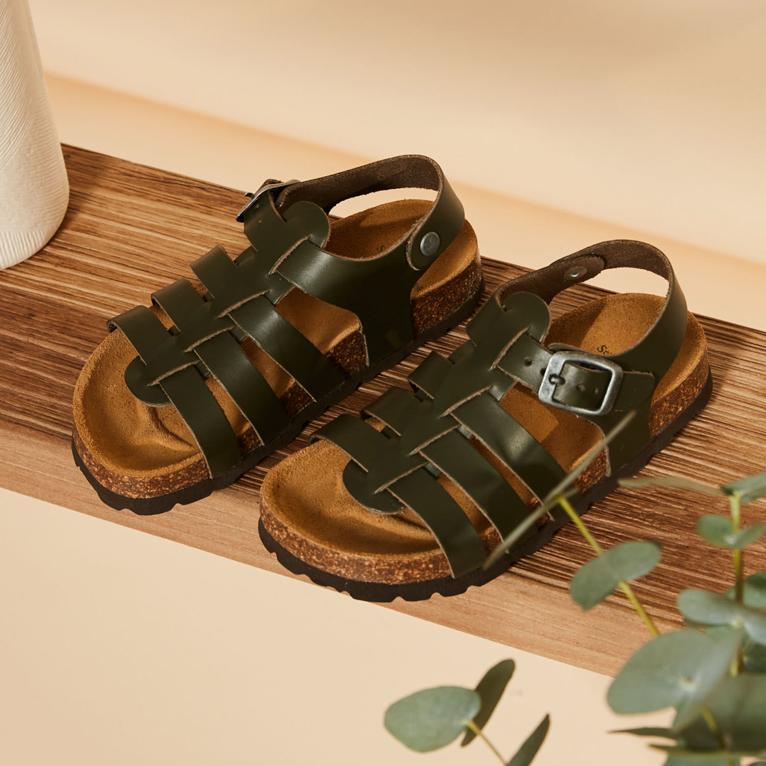 Sandálias verdes escuras de criança com sola castanha e fivela metálica sobre madeira