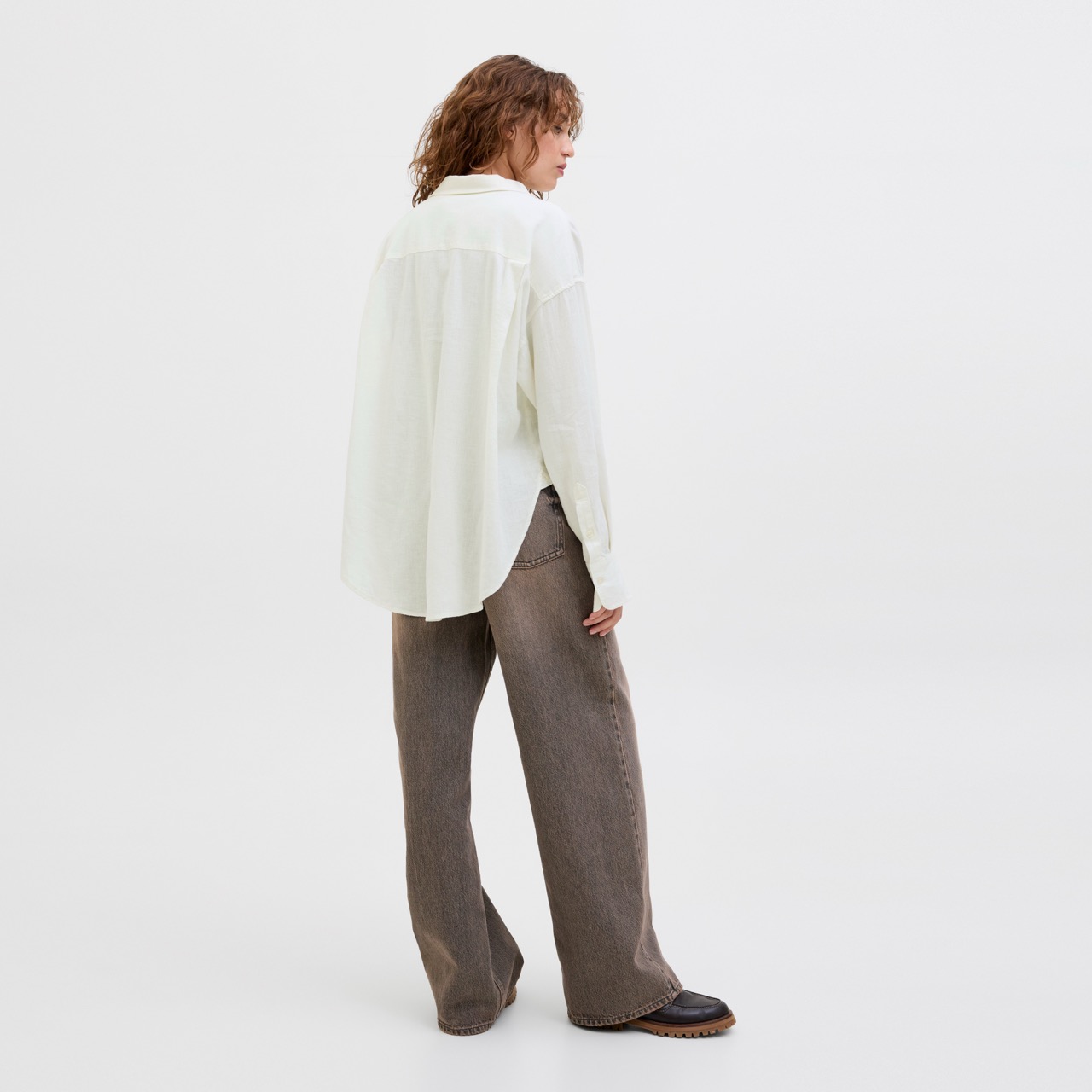 Pessoa de costas com camisa branca e calças castanhas largas em fundo branco