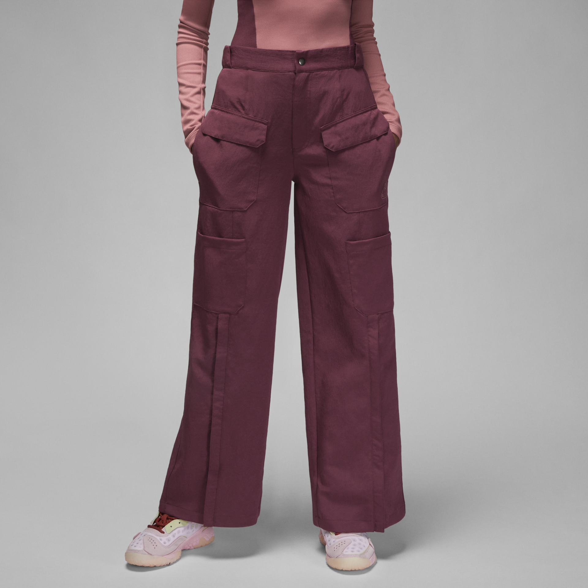 Calças largas bordeaux com bolsos grandes, camisola rosa e ténis rosa claro em fundo cinza.