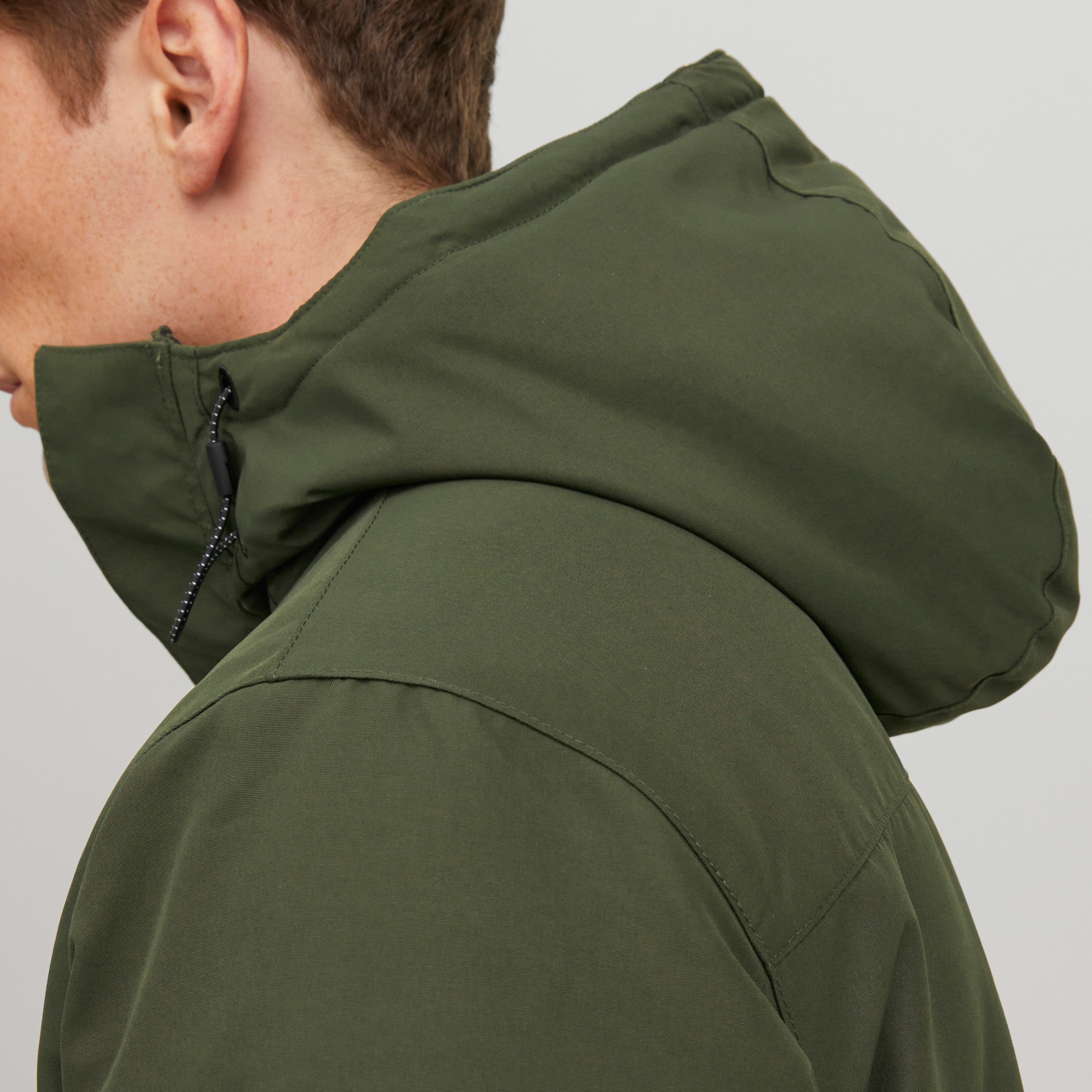 Pessoa com casaco verde-oliva com capuz e fecho de cordão preto, fundo cinza