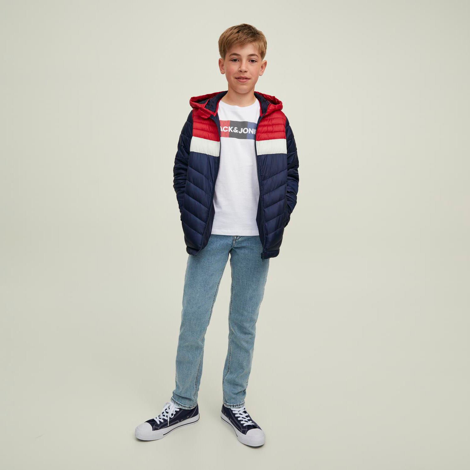 Menino com casaco acolchoado vermelho, branco e azul escuro e camisola branca com padrão colorido em fundo neutro.