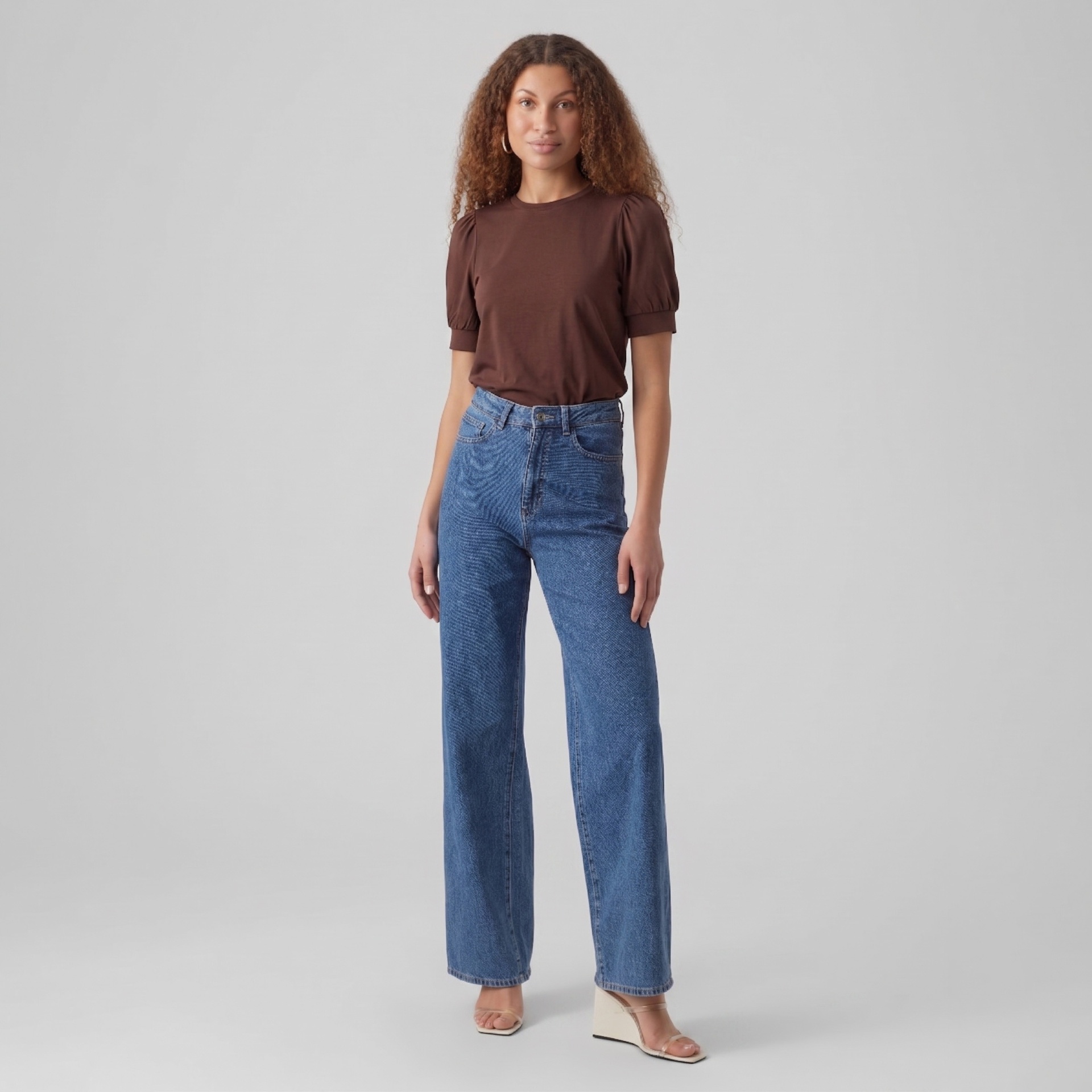 Mulher a usar t-shirt castanha com mangas franzidas, calças jeans azul claro e sandálias bege de cunha num fundo cinzento.