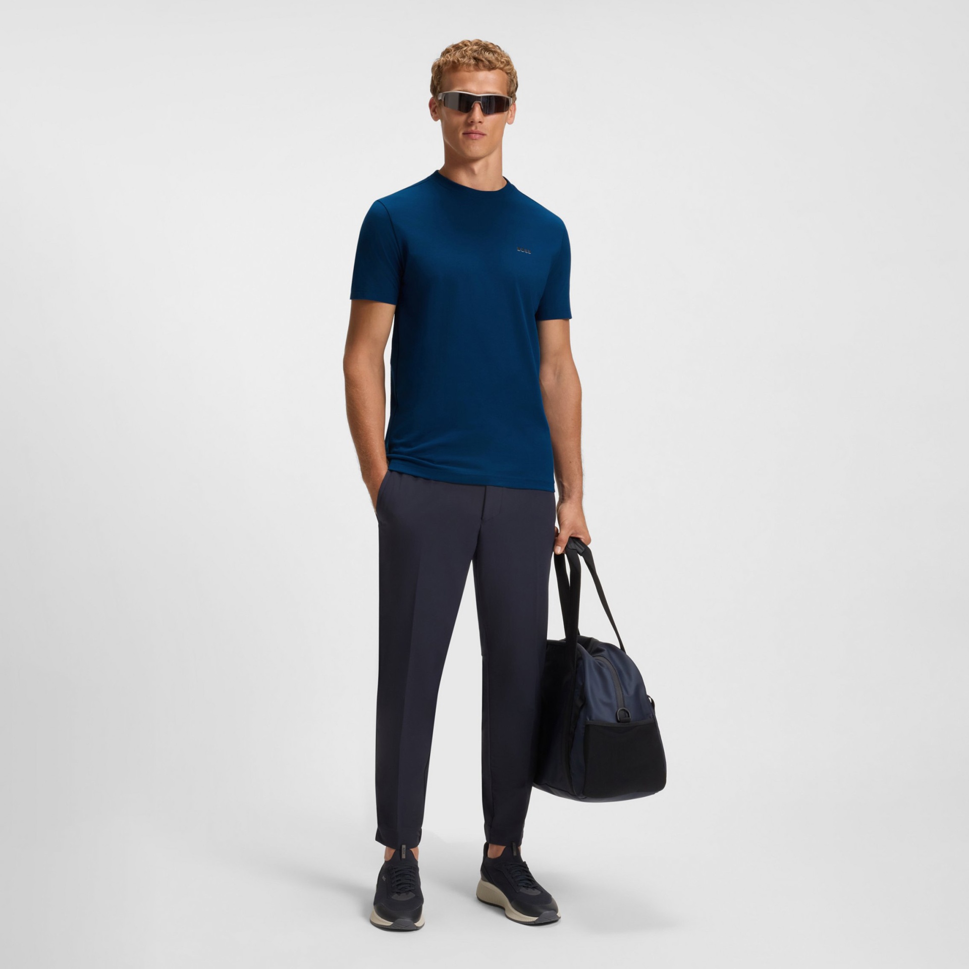 Homem com t-shirt azul escura, calças pretas, sapatos pretos e mala preta.