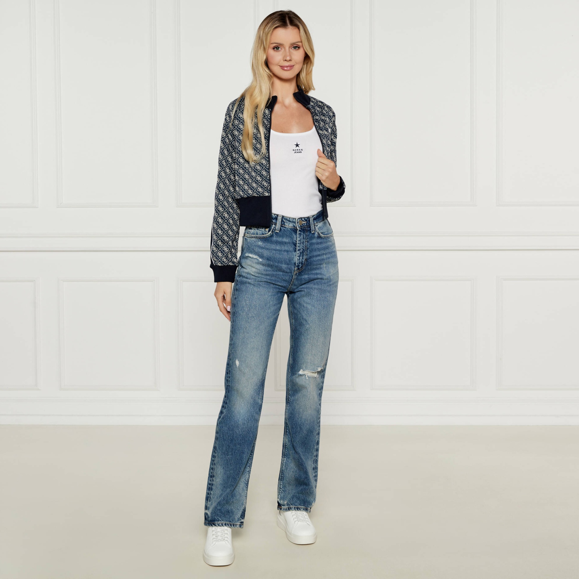 Mulher com casaco azul padrão branco, camisola branca com texto GUESS JEANS, jeans azuis e ténis brancos em fundo branco