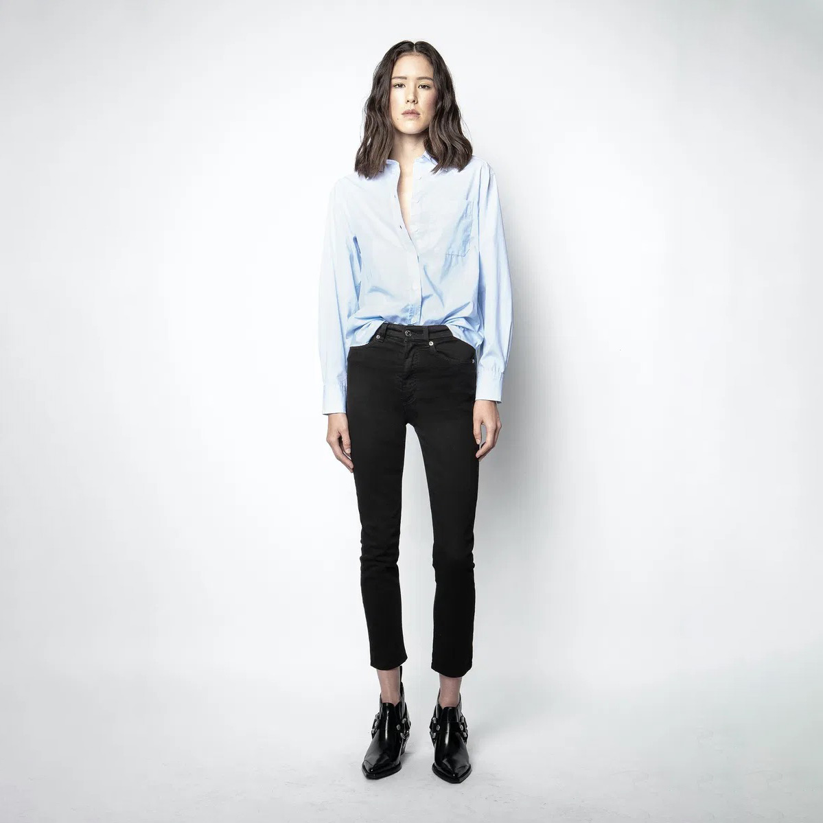 Mulher com camisa azul clara, calças pretas e botins pretos em fundo branco