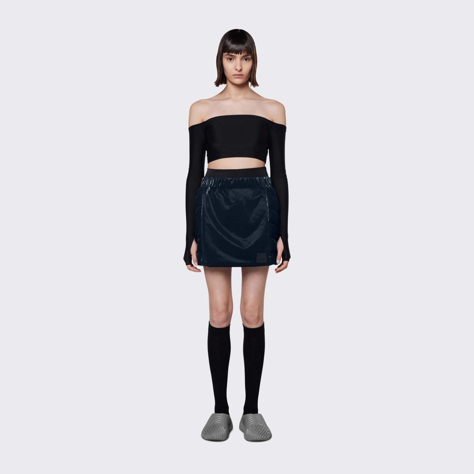 Modelo feminina com top preto ombro a ombro, saia azul escuro, meias pretas e chinelos cinzentos.