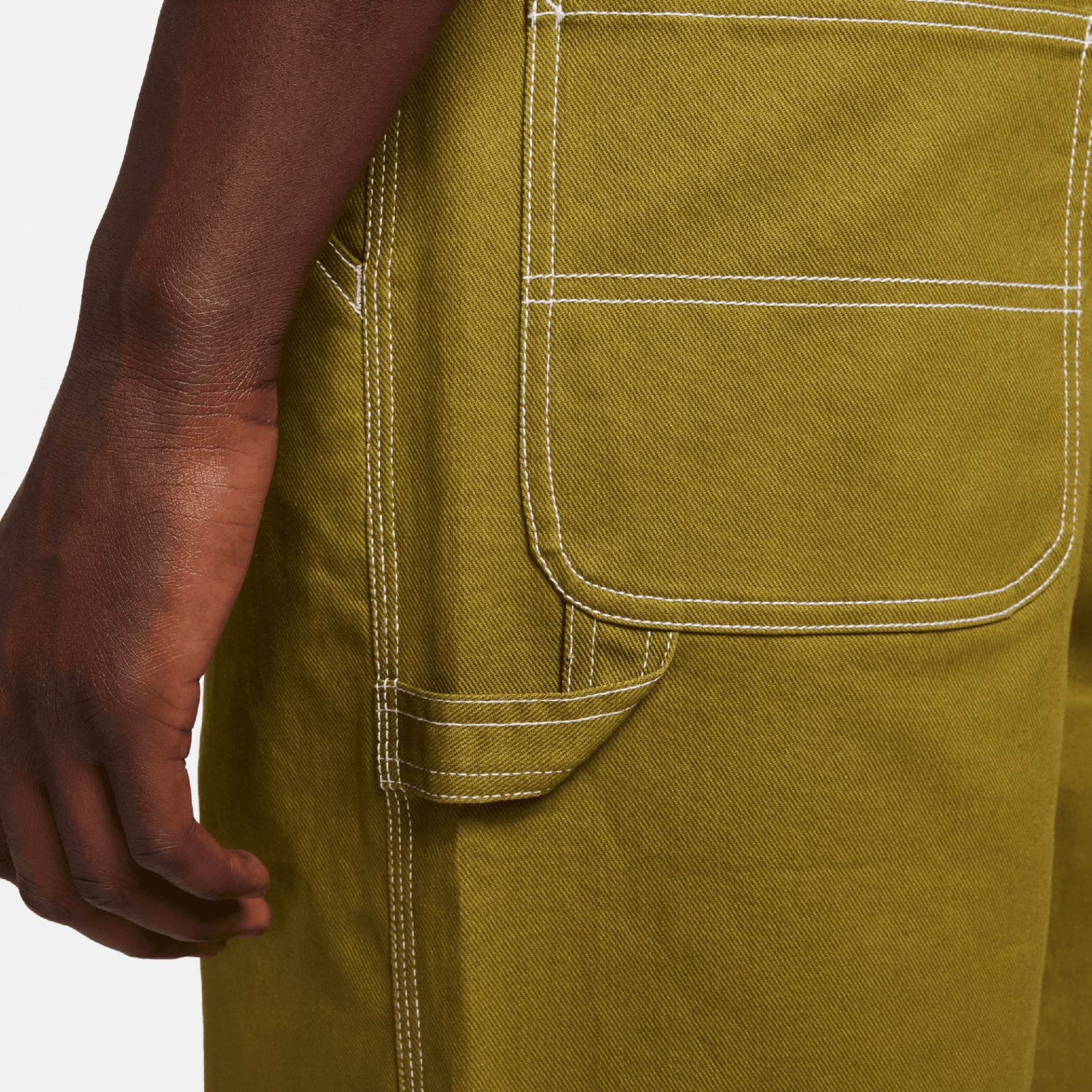 Calças verdes com costuras brancas e bolso grande visível ao lado de uma mão.