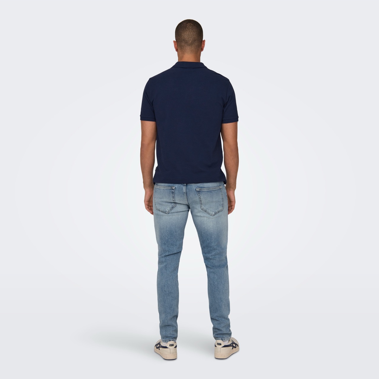Homem de costas com polo azul, jeans azuis claros e ténis brancos