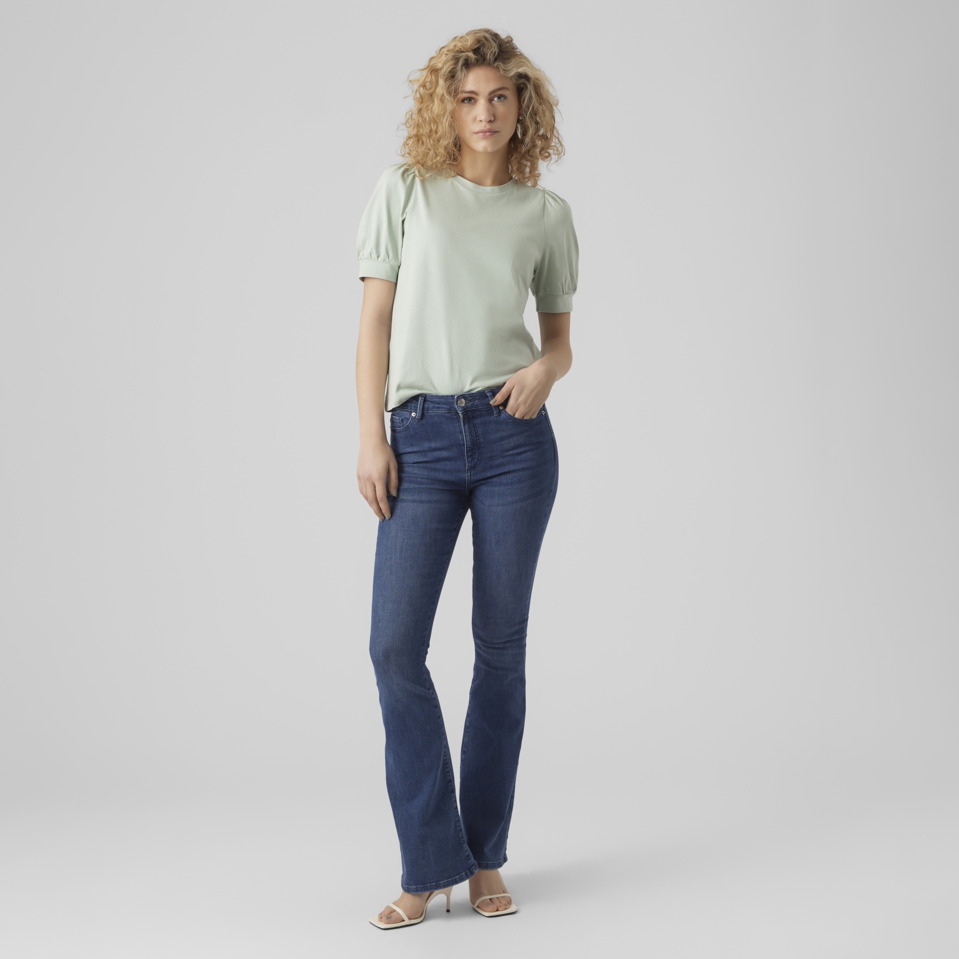Mulher com t-shirt verde claro, jeans azul escuro e sapatos bege em fundo cinza