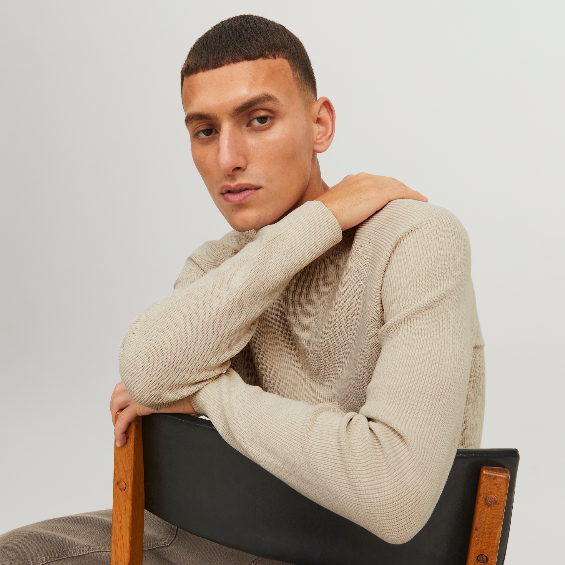 Homem com camisola bege de malha fina sentado numa cadeira de madeira com encosto preto