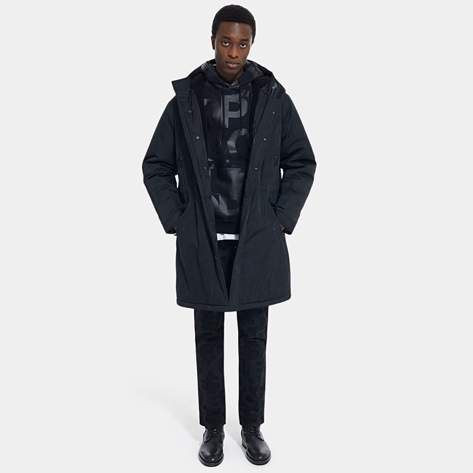 Homem com casaco comprido preto, sweatshirt estampada, calças pretas estampadas e sapatos pretos numa sala branca