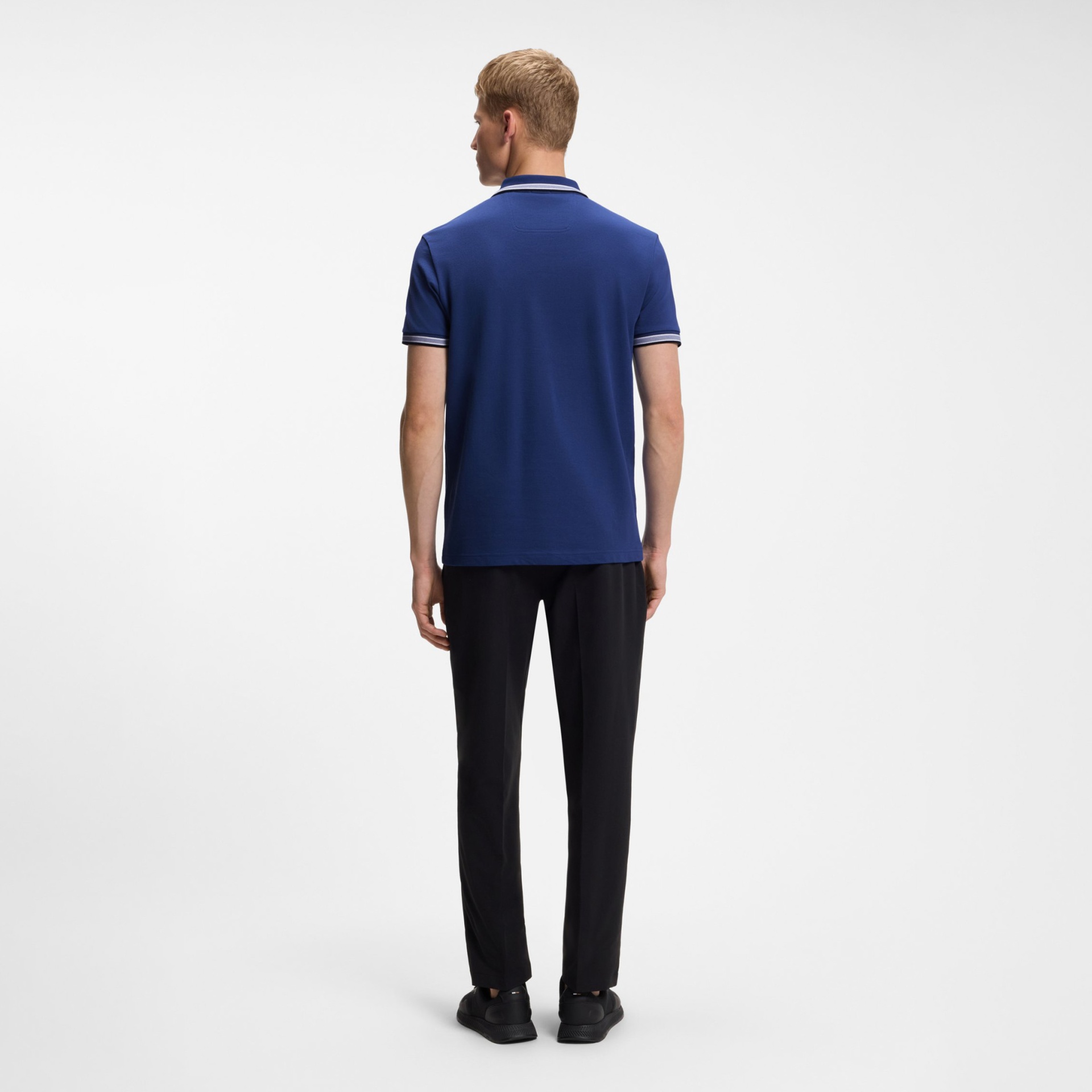 Homem de costas com polo azul, calças pretas e sapatos pretos sobre fundo branco.