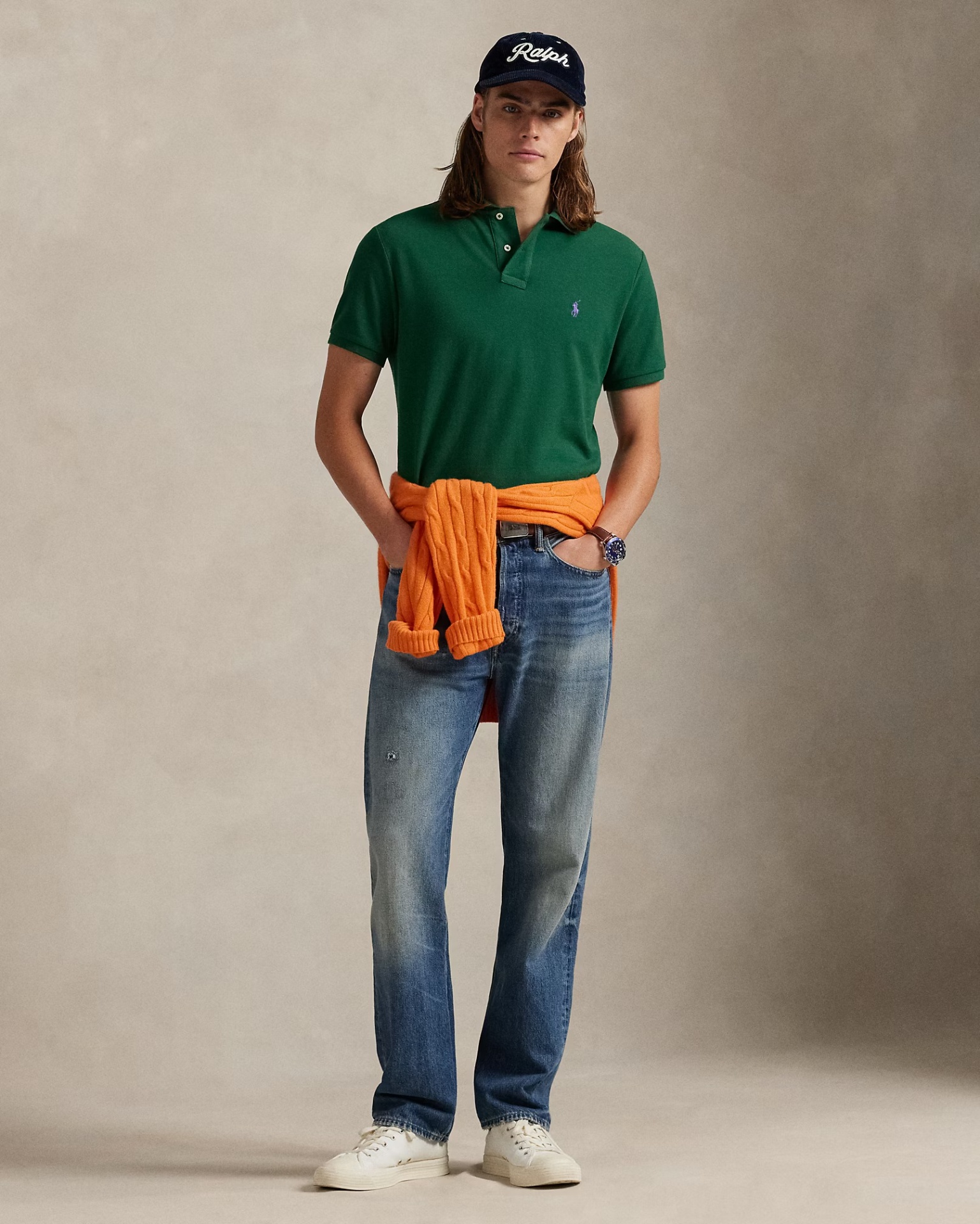 Homem com polo verde, jeans azuis, casaco laranja atado à cintura, sapatilhas brancas e boné preto com Ralph.