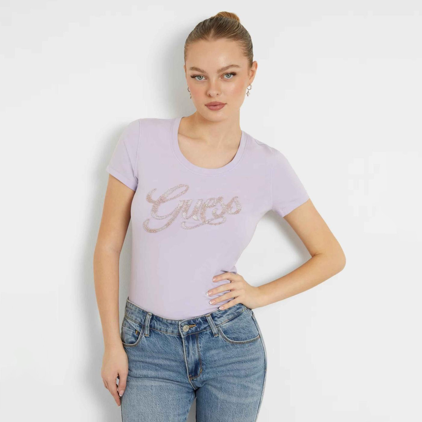 Mulher com t-shirt lilás com logo Guess em strass e jeans azul claro.