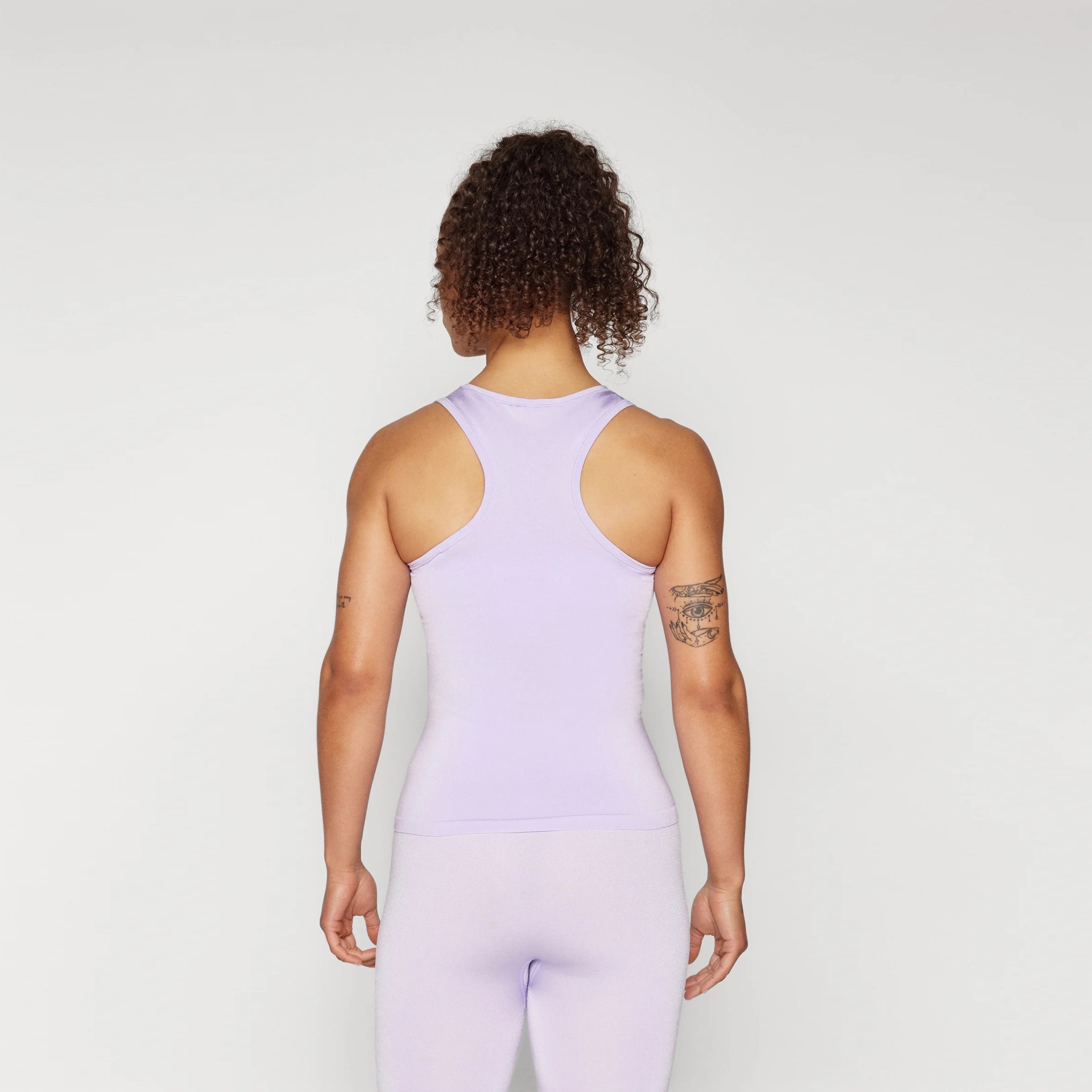 Mulher de costas a usar top e leggings lilás sobre fundo branco