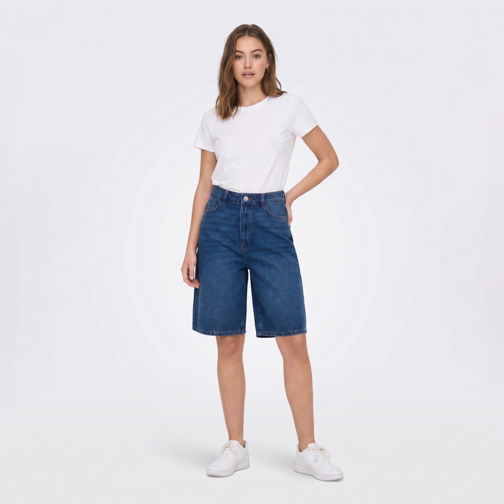 Modelo feminina com t-shirt branca, calções de ganga azuis e ténis brancos em fundo branco