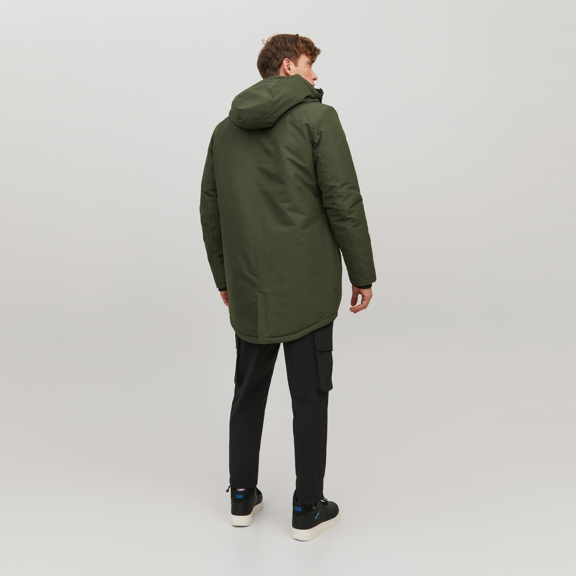Homem de costas a usar casaco verde-oliva com capuz, calças pretas e sapatilhas pretas com sola branca e texto azul JACK & JONES.