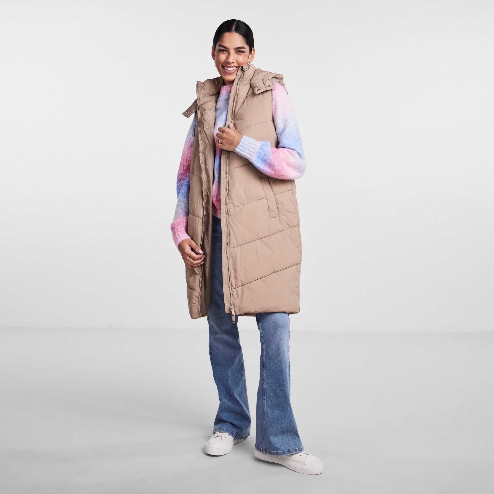 Mulher sorridente com colete comprido bege, camisola multicolorida, jeans azuis e sapatilhas brancas.