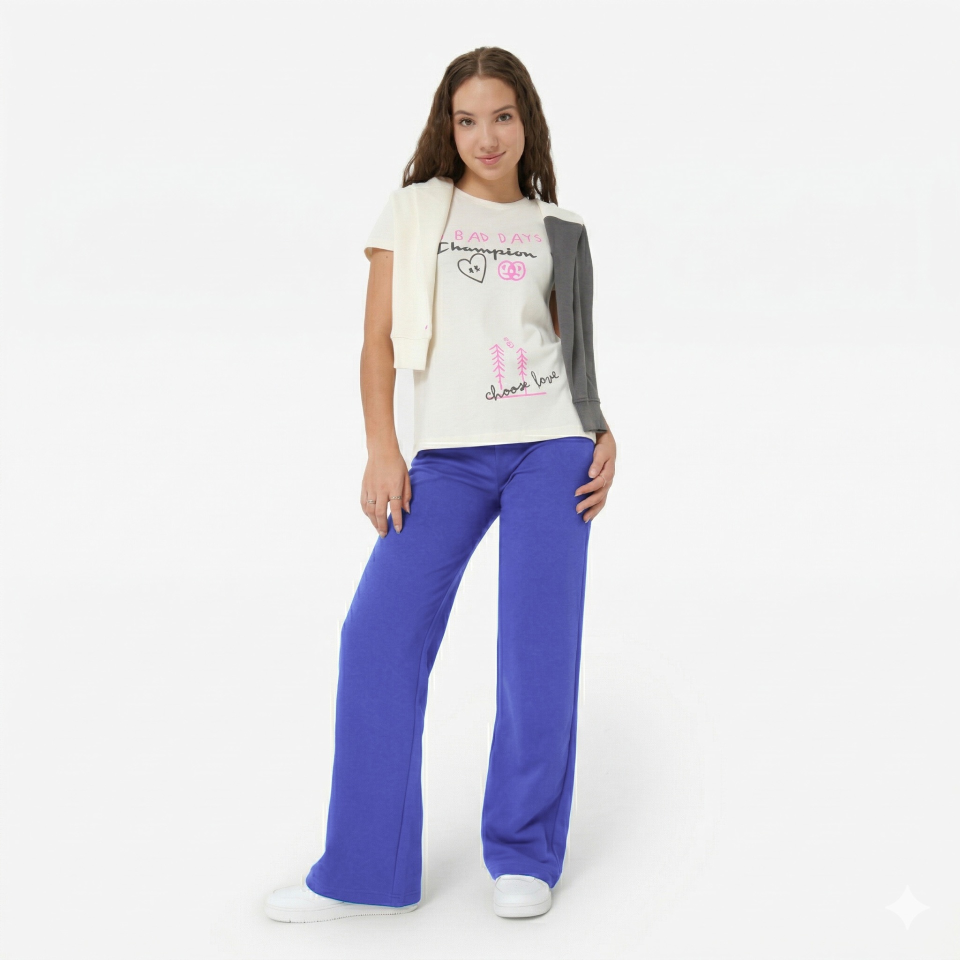 Pessoa jovem com roupa casual: t-shirt branca estampada, calças largas azuis e ténis brancos, branco limpo ao fundo.