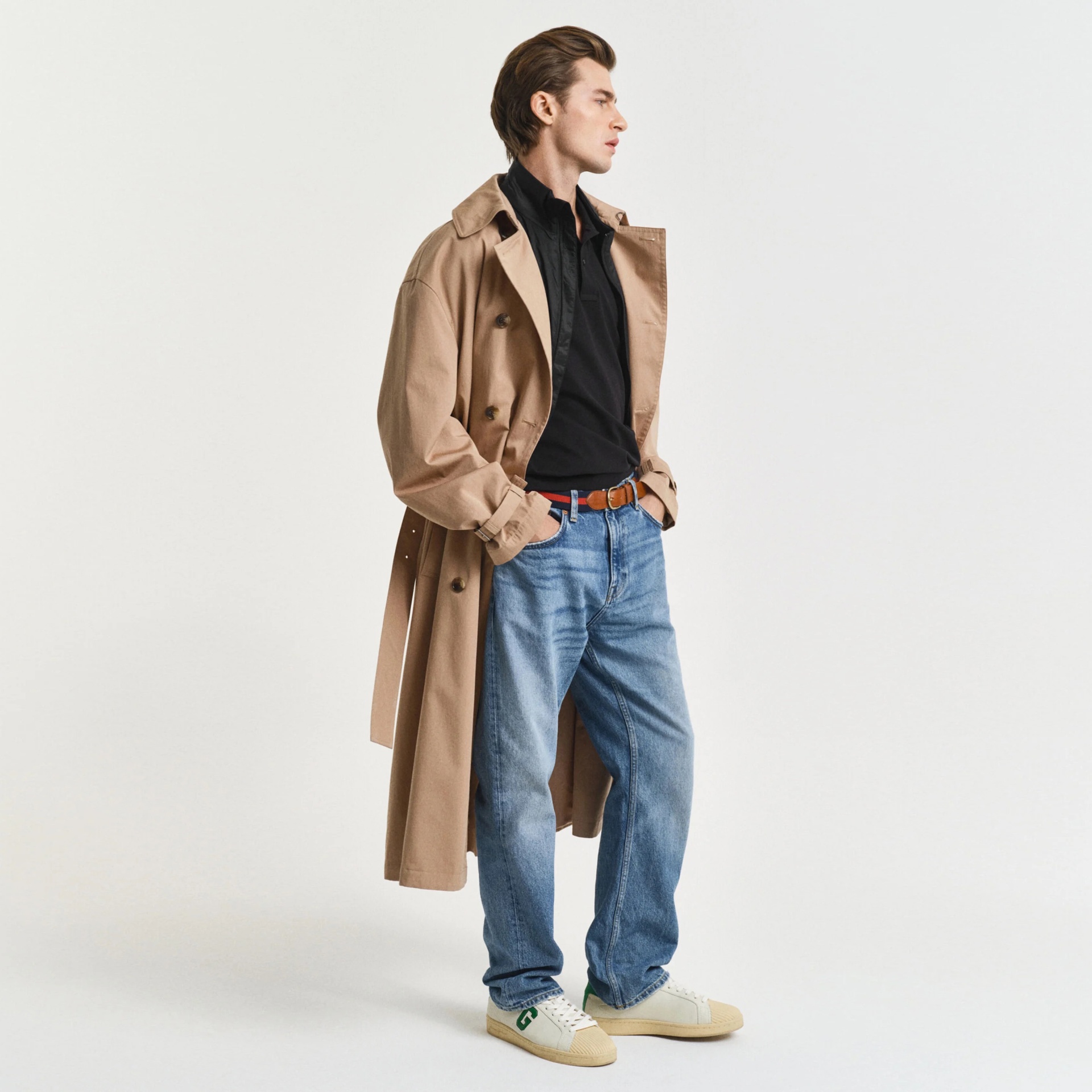 Homem com casaco trench coat bege, camisa preta, jeans azuis, cinto castanho e ténis brancos.