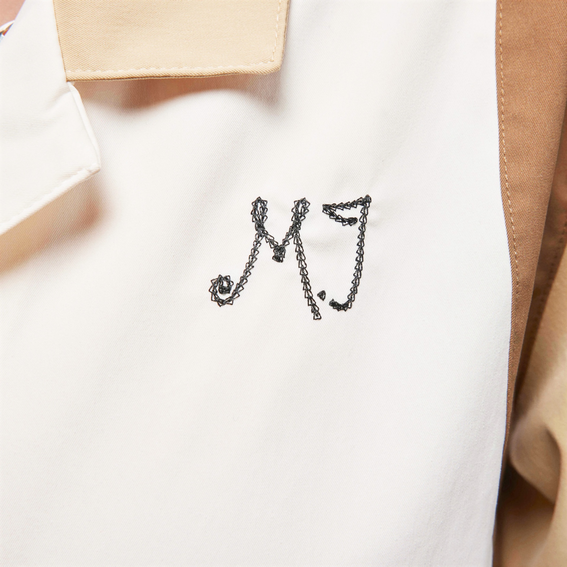 Peça de roupa branca e bege com bordado preto das letras M e J.
