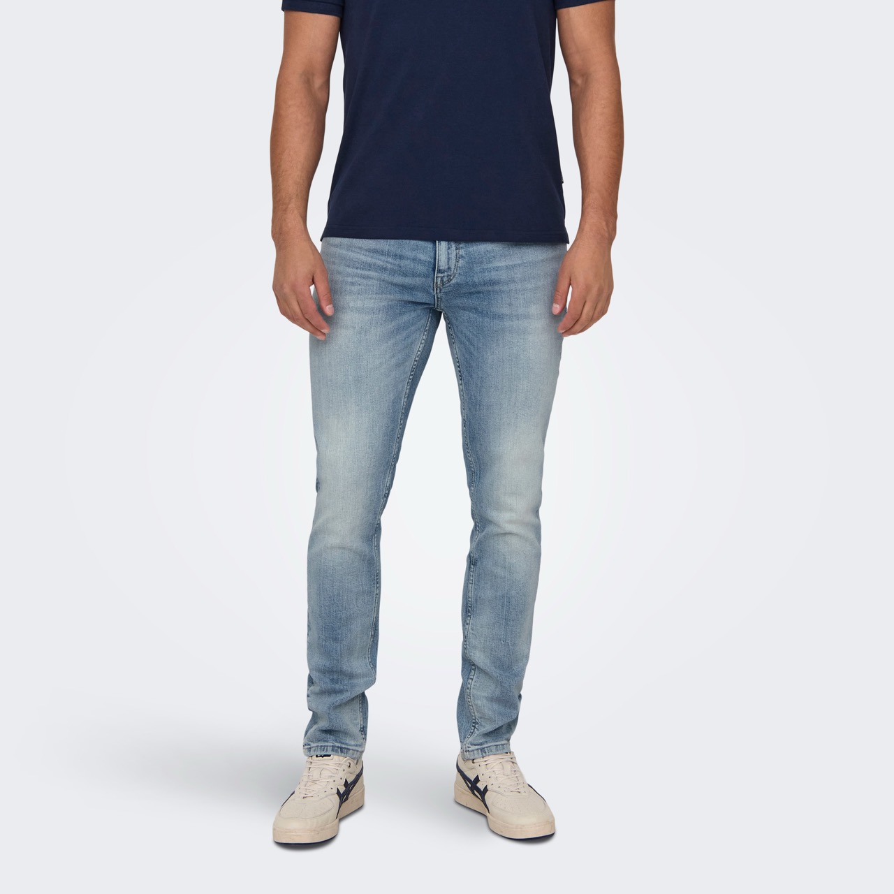 Homem vestindo polo azul escuro, jeans claro desbotados, e ténis brancos com detalhes azuis