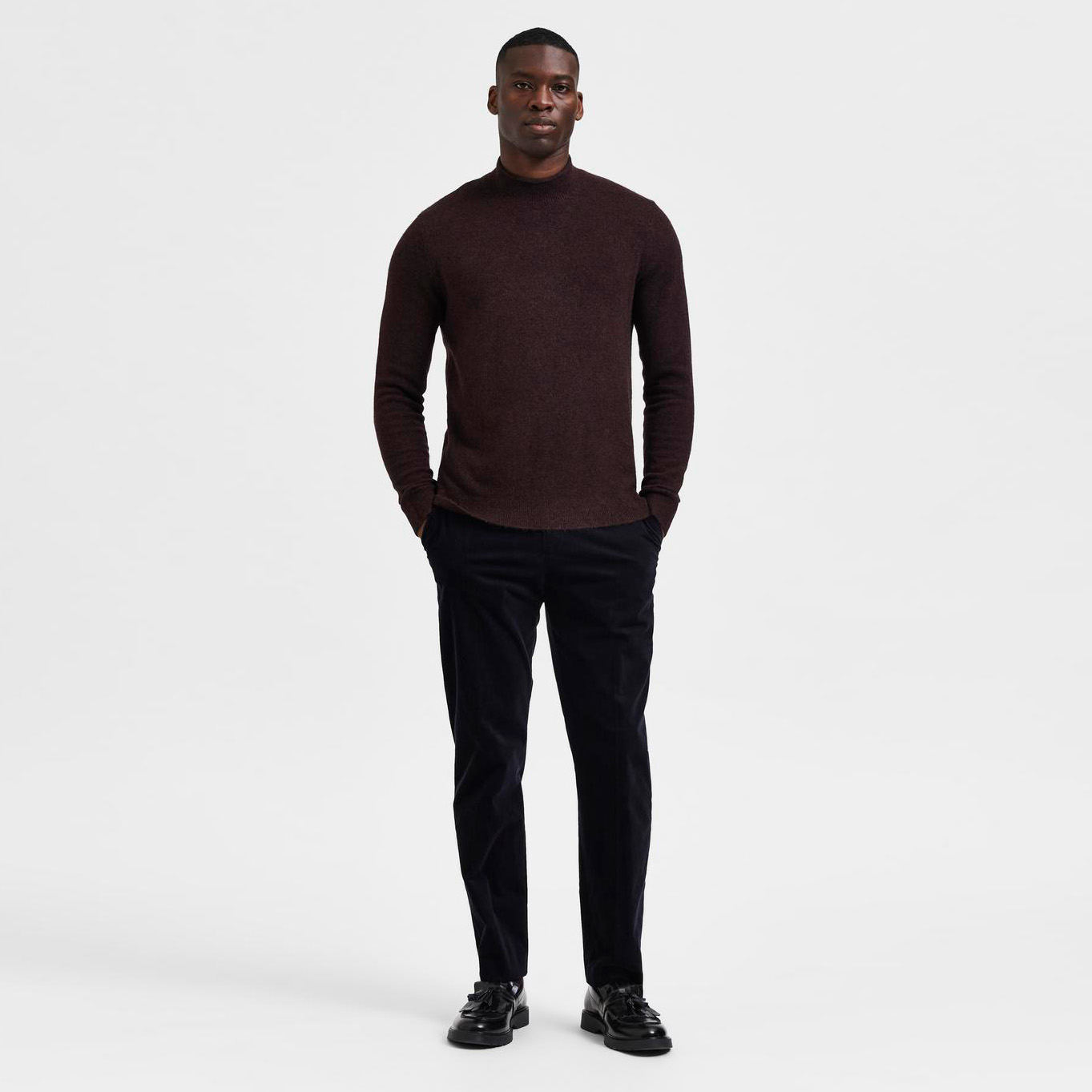 Homem de pé com camisola castanha de gola alta e calças pretas