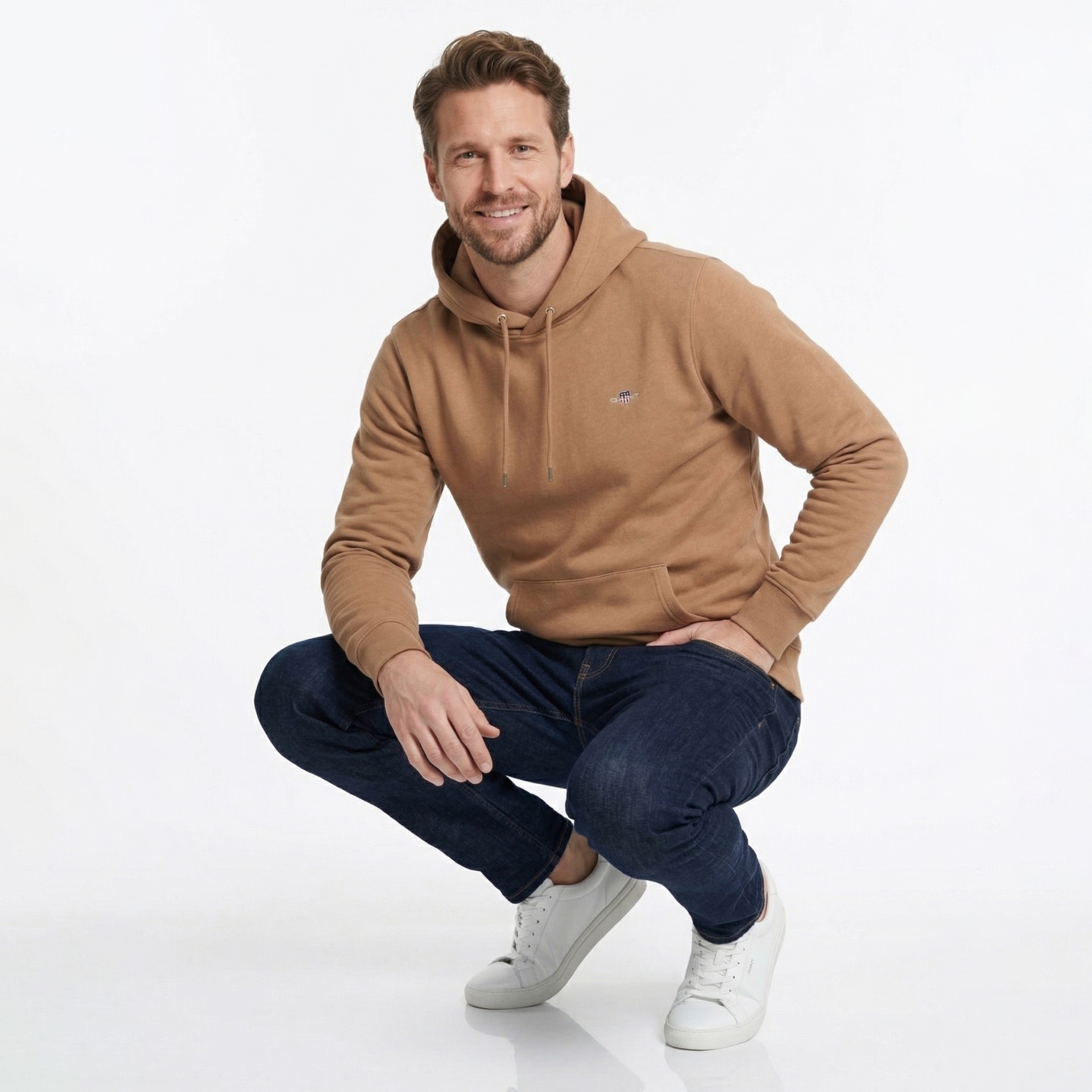 Homem de agachado com capuz castanho, jeans azuis e ténis brancos em fundo branco