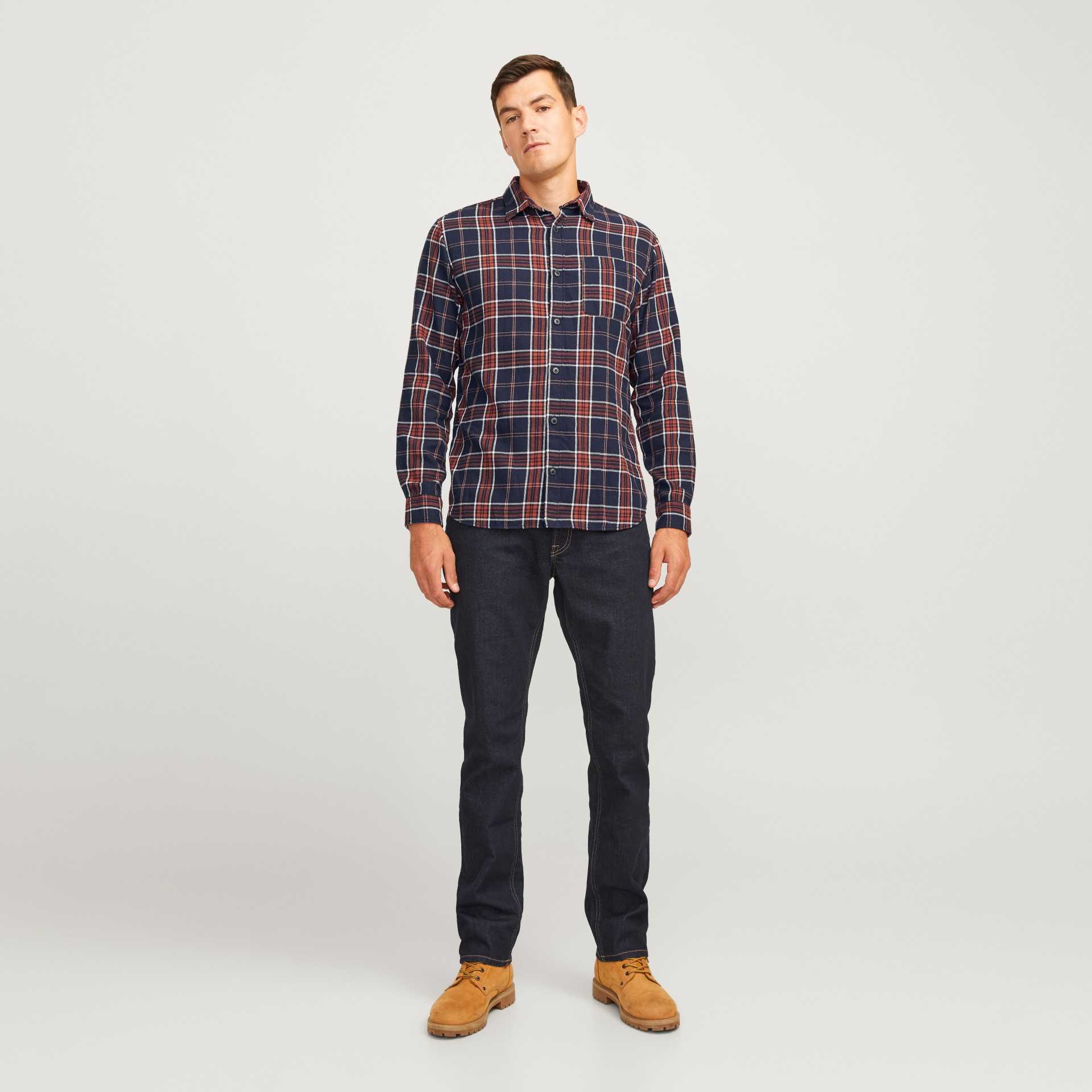 Homem com camisa aos quadrados, calças castanhas escuras e botas castanhas em fundo branco