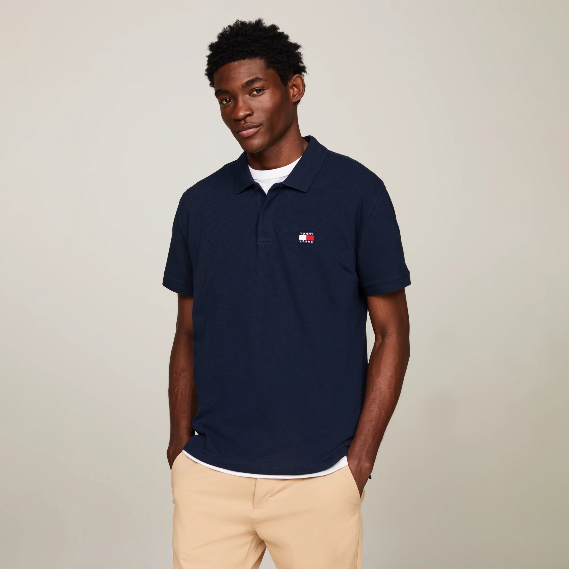 Homem a usar polo azul escura e calças bege com fundo bege claro