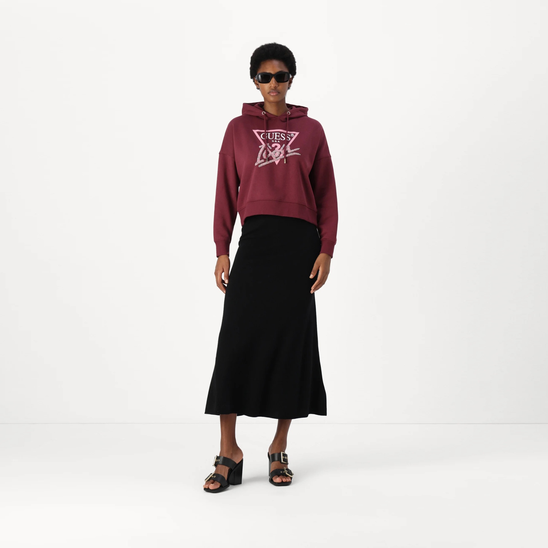 Mulher com hoodie bordô GUESS, saia preta longa, sapatos de salto pretos e óculos escuros.