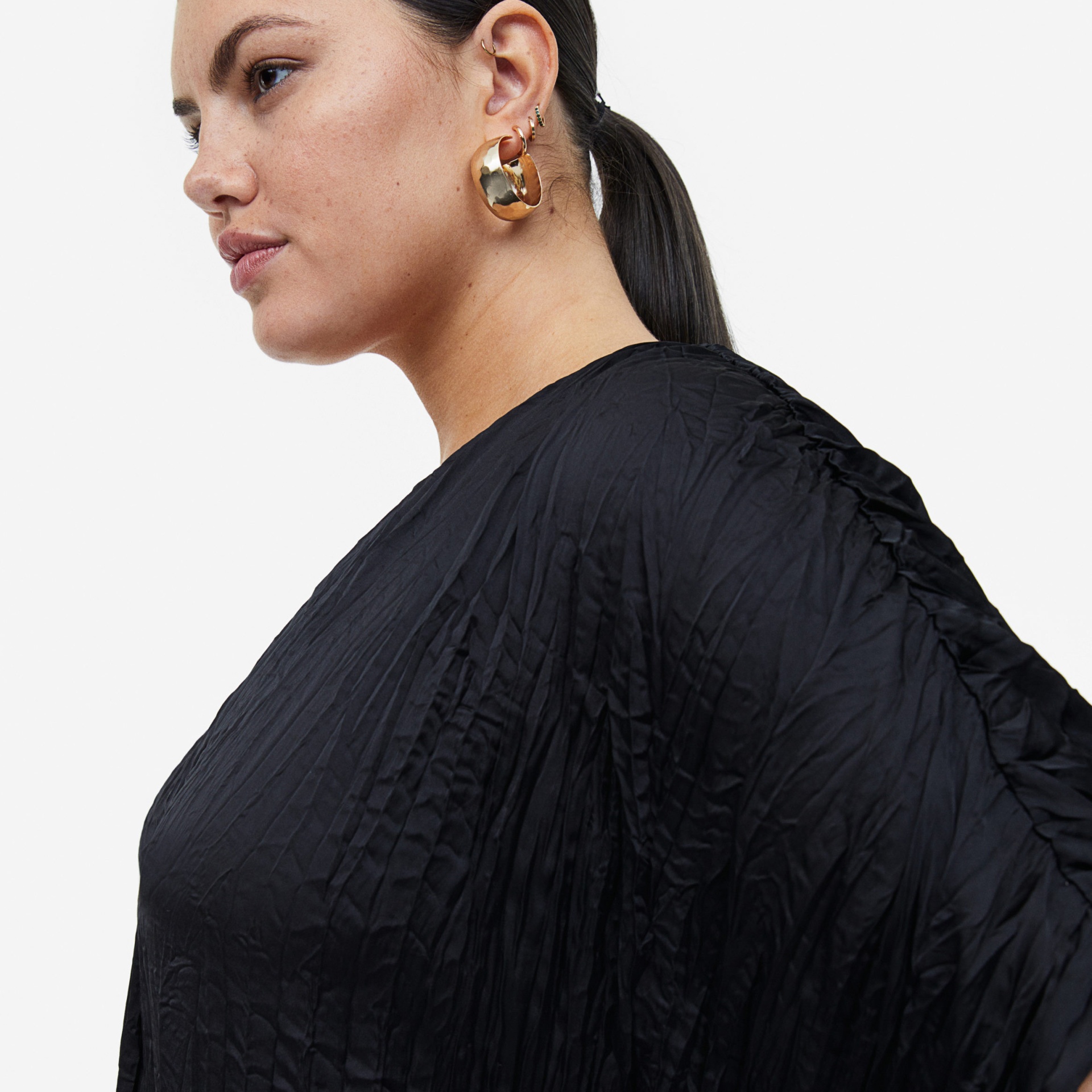 Pessoa com pendente e argolas douradas, camisa preta enrugada, cabelo preto apanhado.