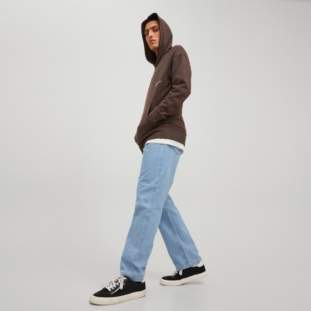 Homem vestido com hoodie castanho, calças de ganga azul claro e ténis pretos com sola branca em fundo neutro