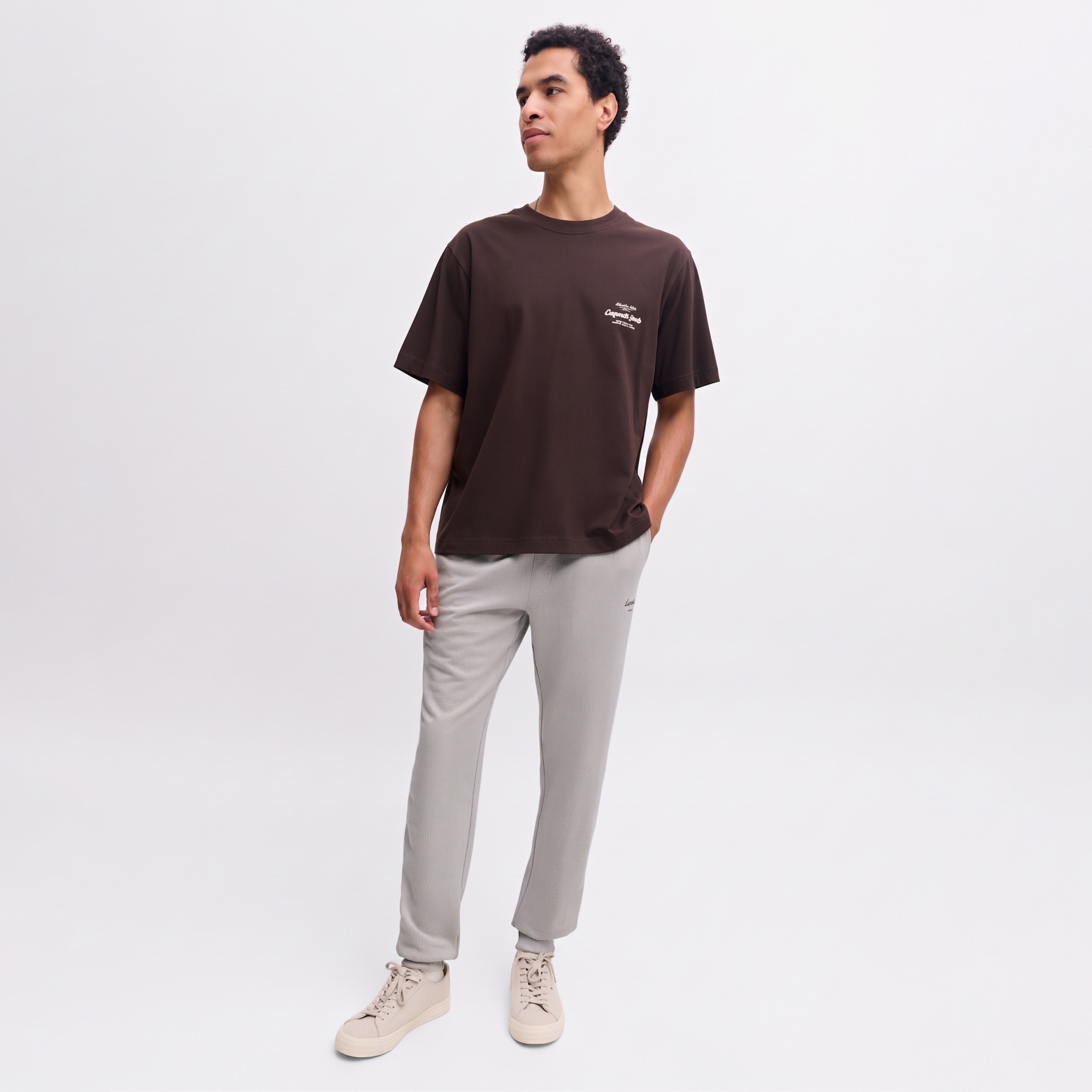 Homem a vestir t-shirt castanha com texto branco, calças cinza claras e sapatilhas bege