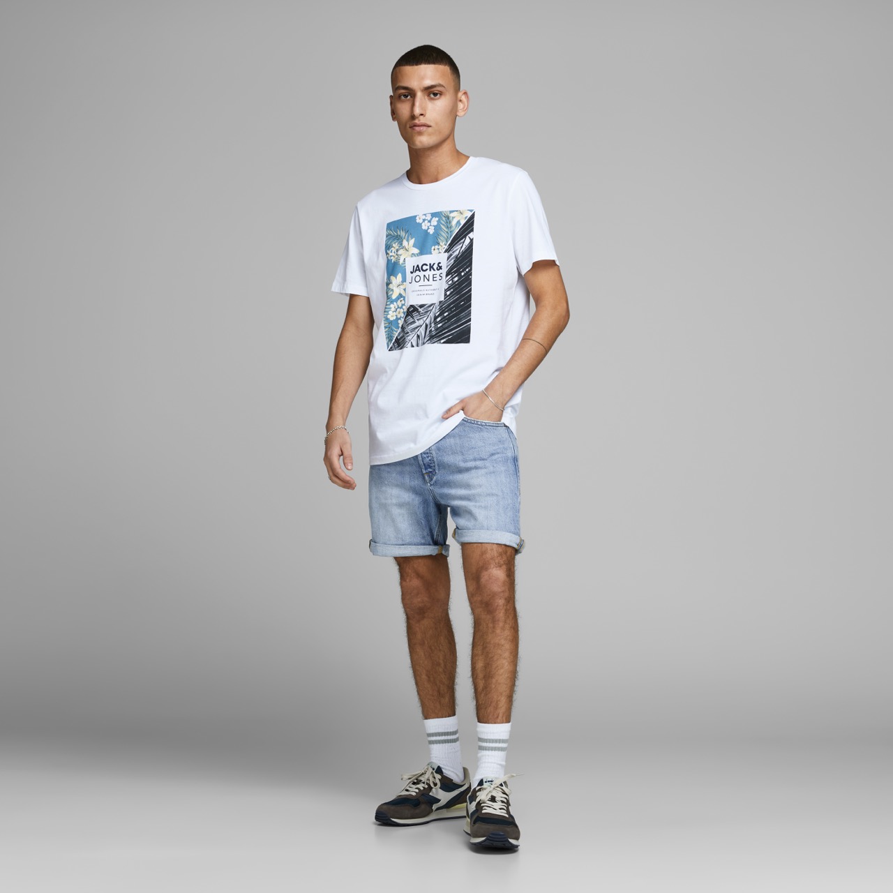 Homem de t-shirt branca estampada, calções de ganga, meias brancas e sapatilhas em fundo cinza