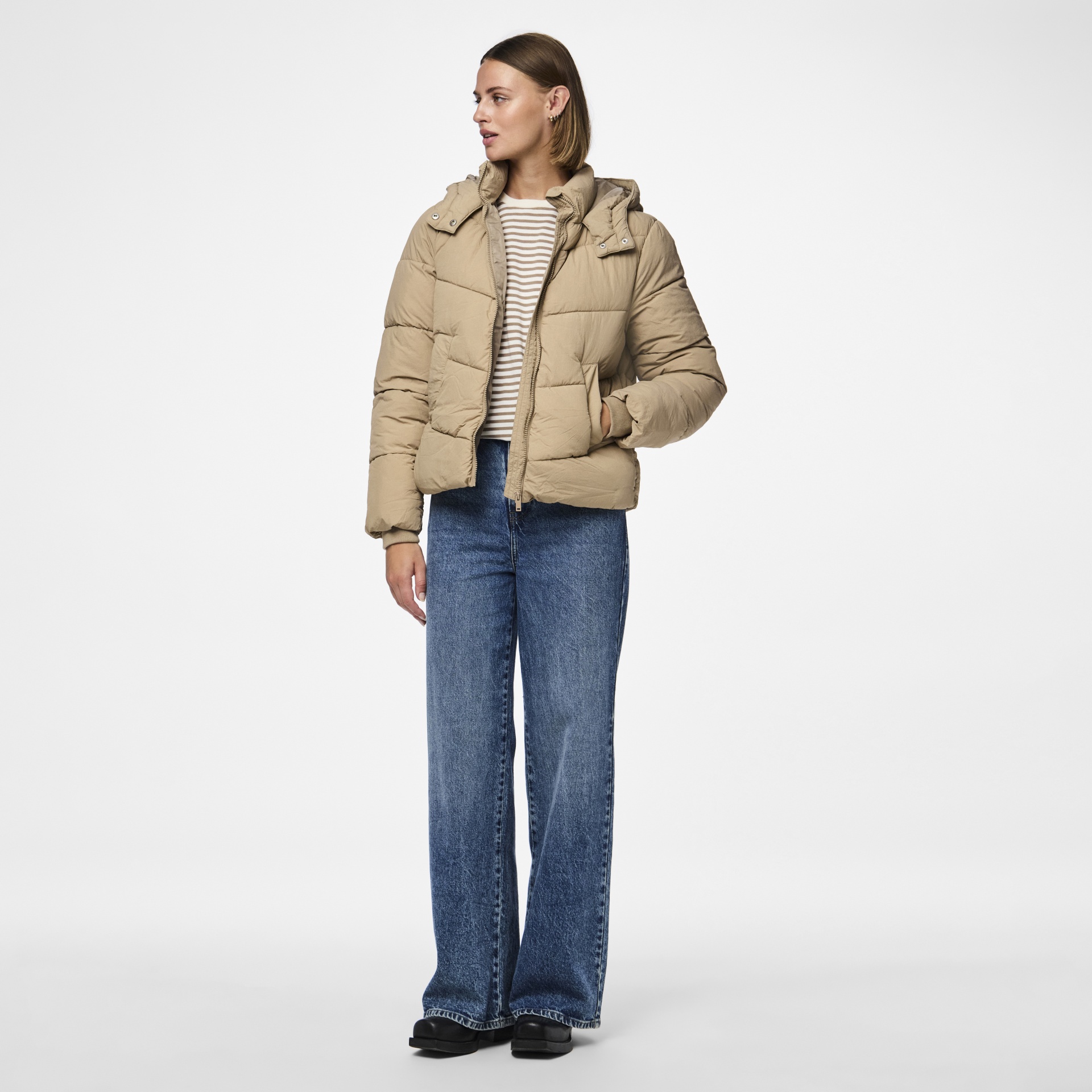 Pessoa com casaco acolchoado bege, camisola riscada, calças de ganga azuis e sapatos pretos.