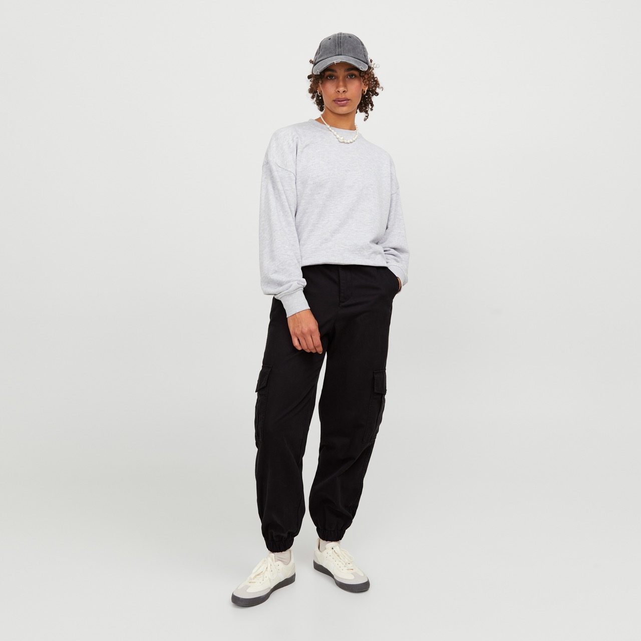 Modelo veste camisola cinzenta, calças cargo pretas, ténis brancos e boné cinzento num fundo branco