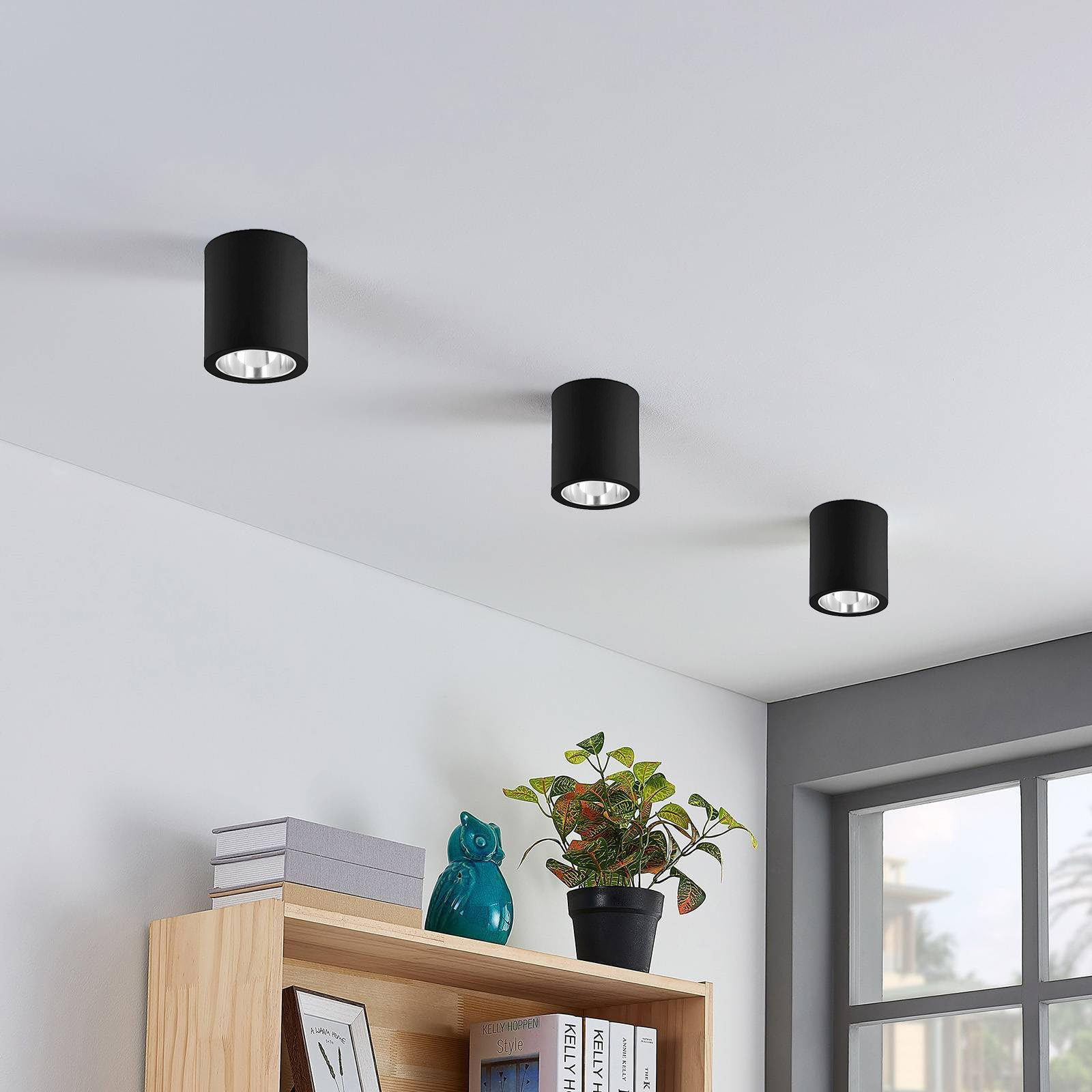três candeeiros de teto pretos cilíndricos num teto branco, estante de madeira com livros, urna azul, planta em vaso preto e janela cinzenta