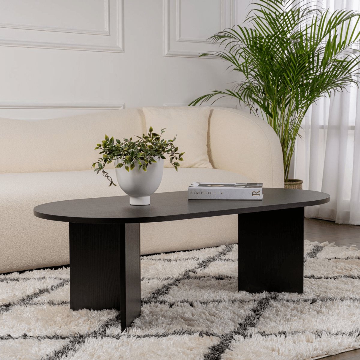 Mesa de centro ovalada preta com vaso e livro, sofá e planta ao fundo