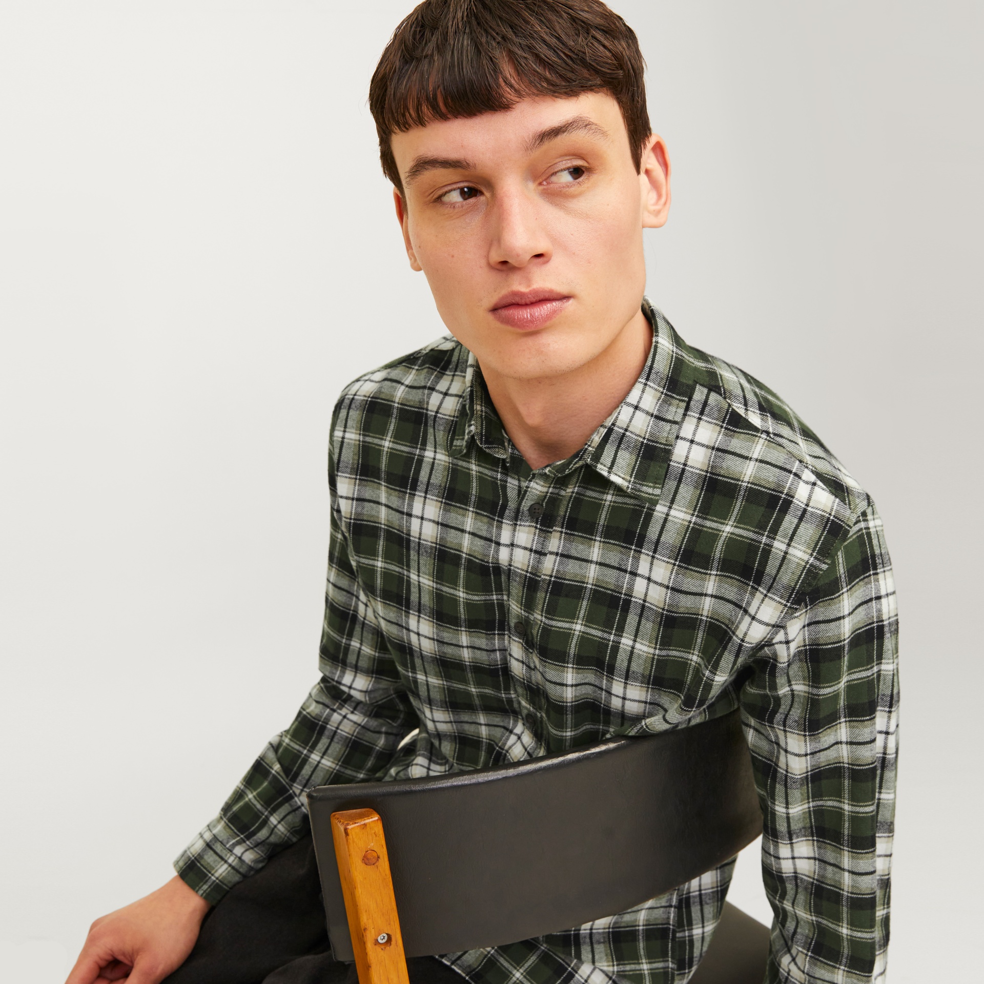Homem com camisa xadrez verde, preta e branca sentado numa cadeira preta com madeira.