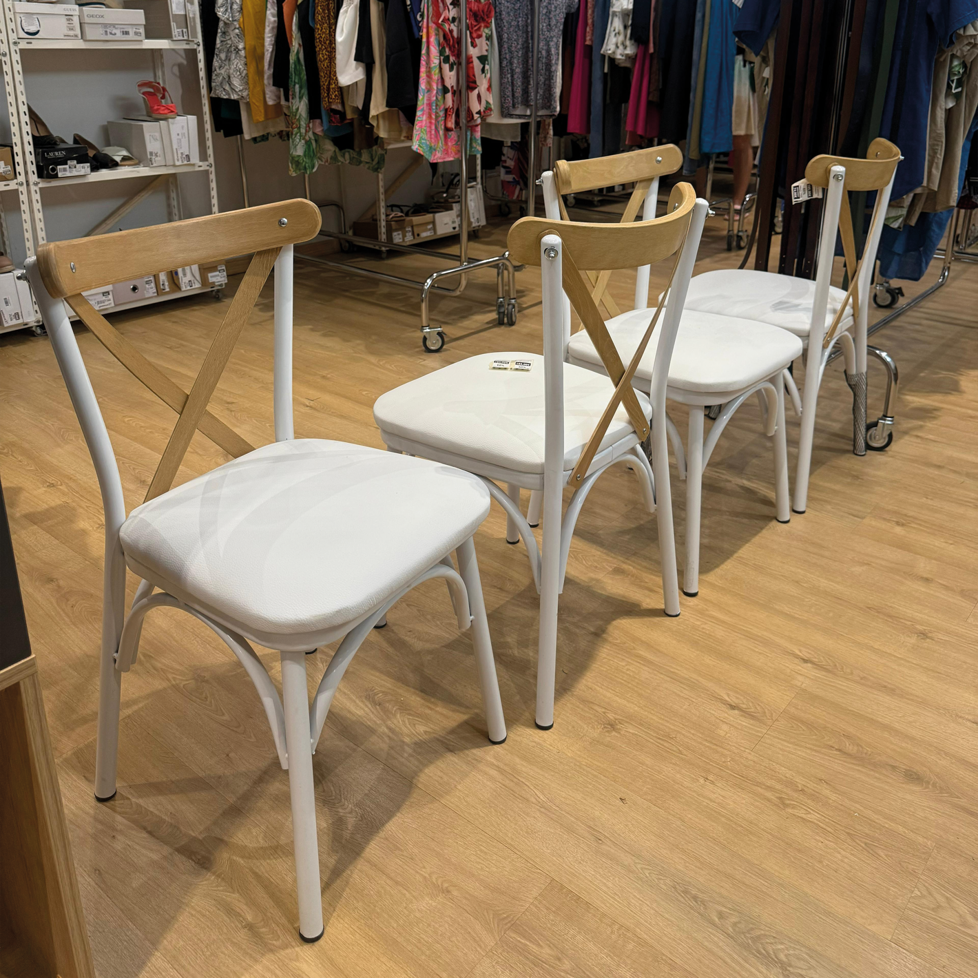 Quatro cadeiras com estrutura branca, encosto em madeira clara e assentos brancos numa loja de roupas