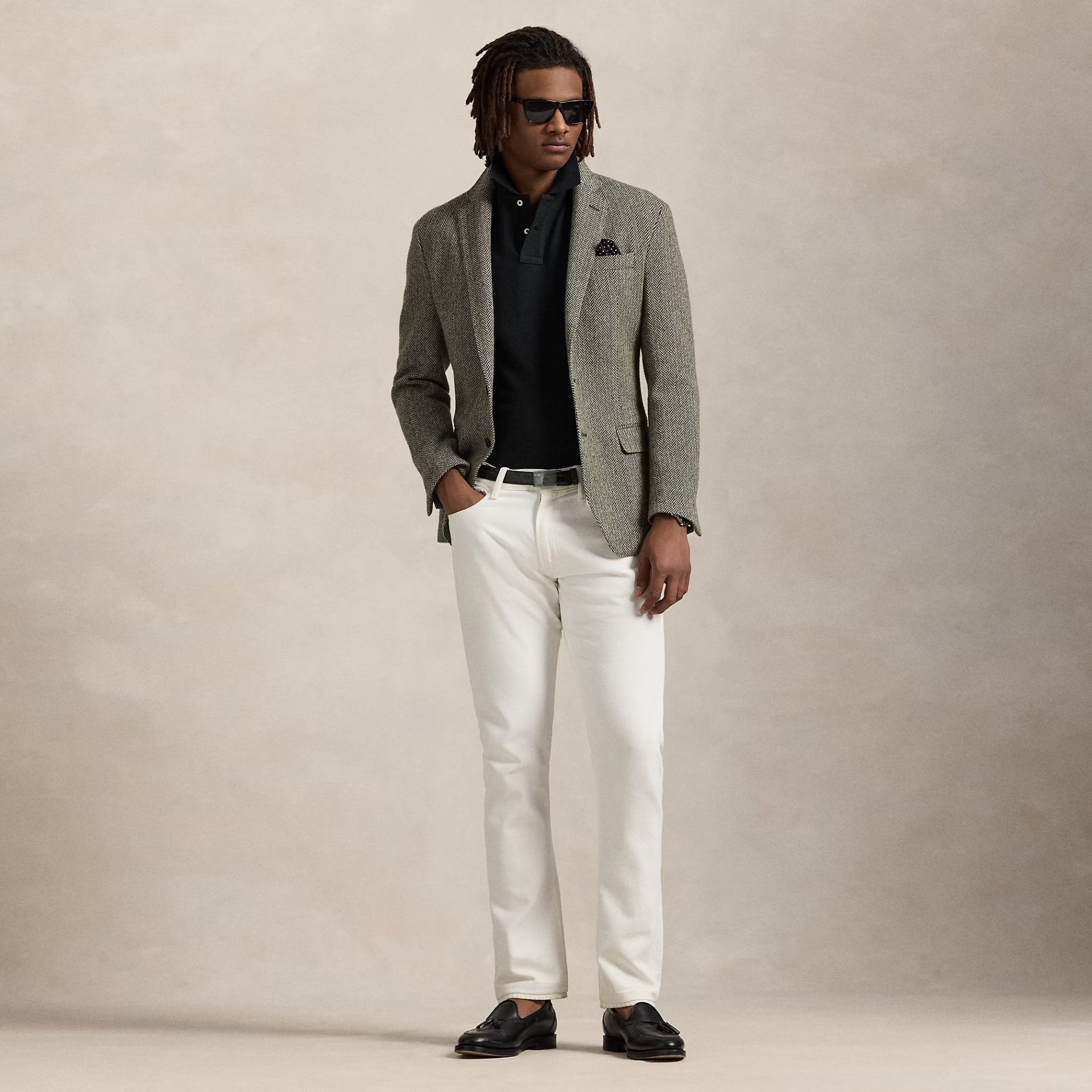 Homem de pé com blazer cinzento, polo preto e calças brancas