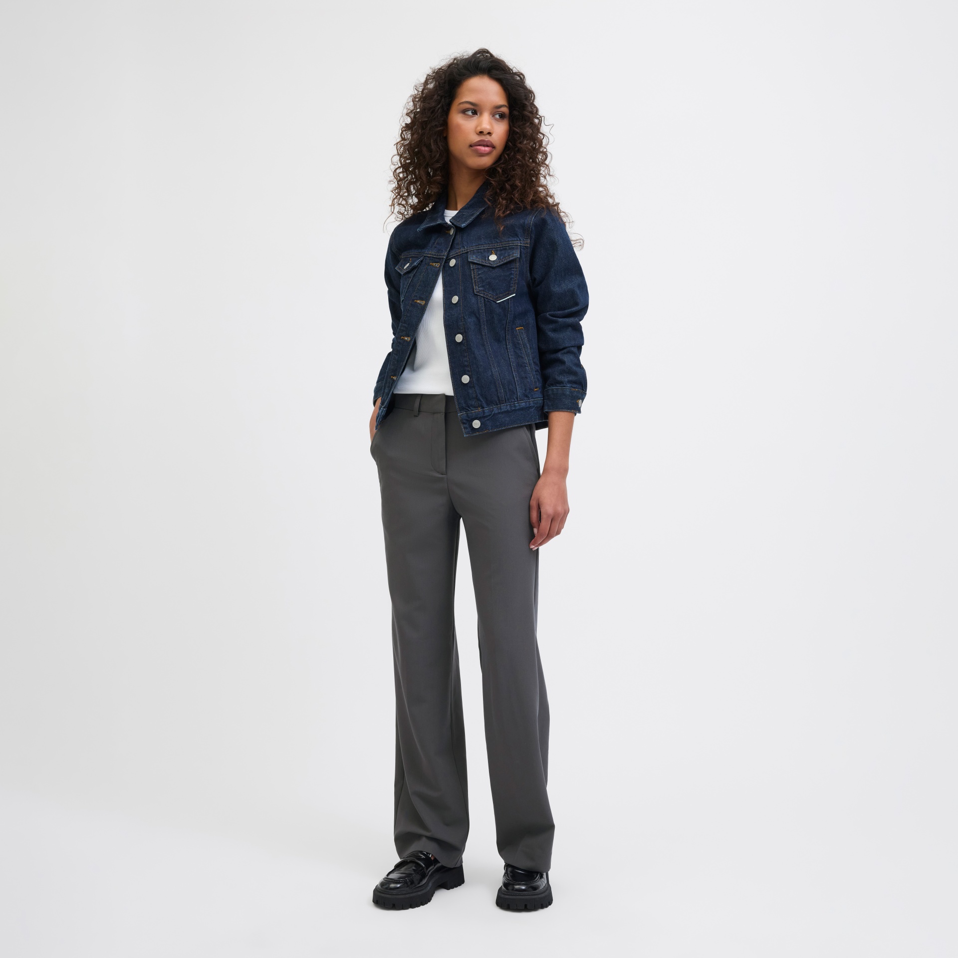 Mulher a usar casaco de ganga azul escuro, camisola branca, calças cinzentas largas e sapatos pretos