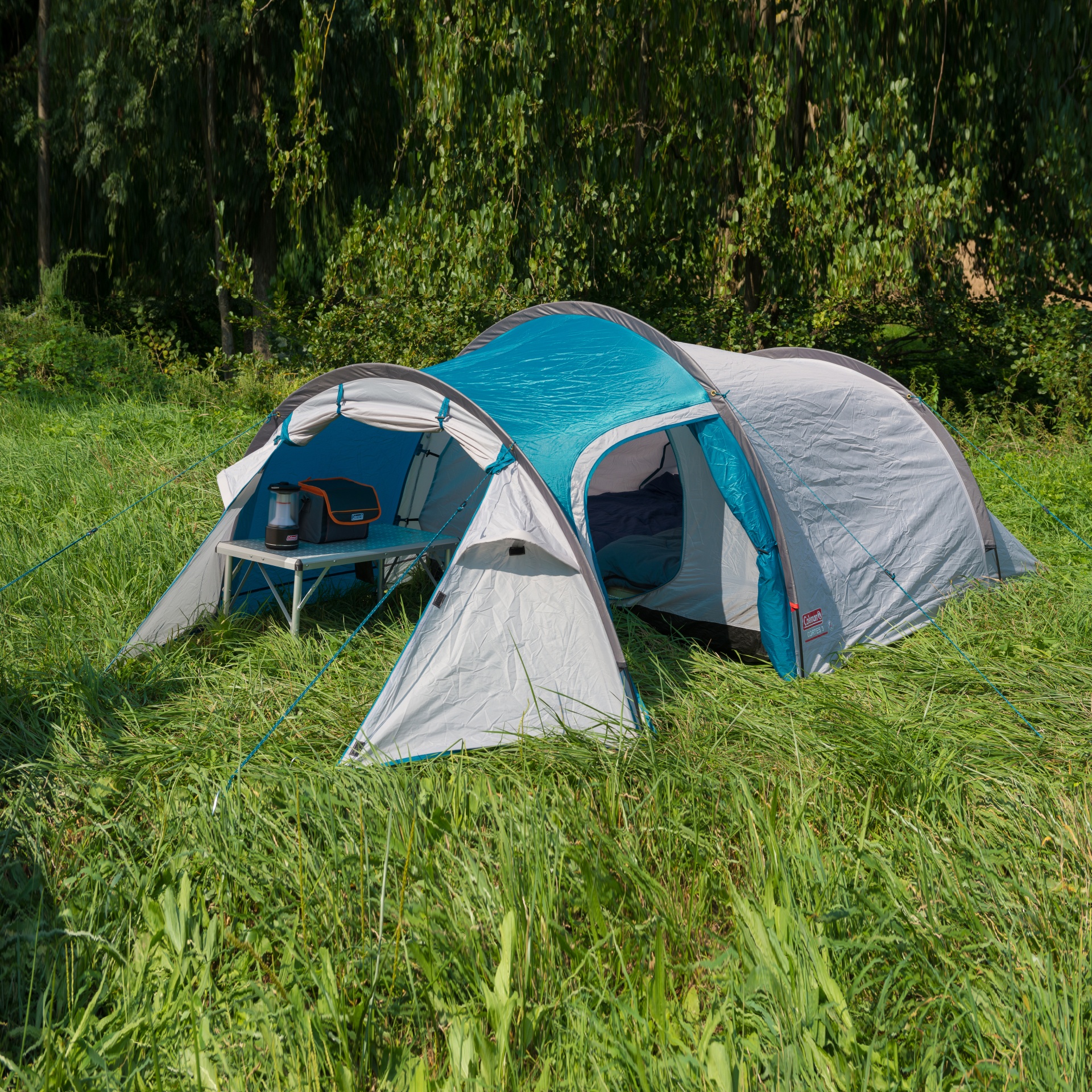 Tenda de campismo azul e cinzenta montada em relva com mesa e lanterna.