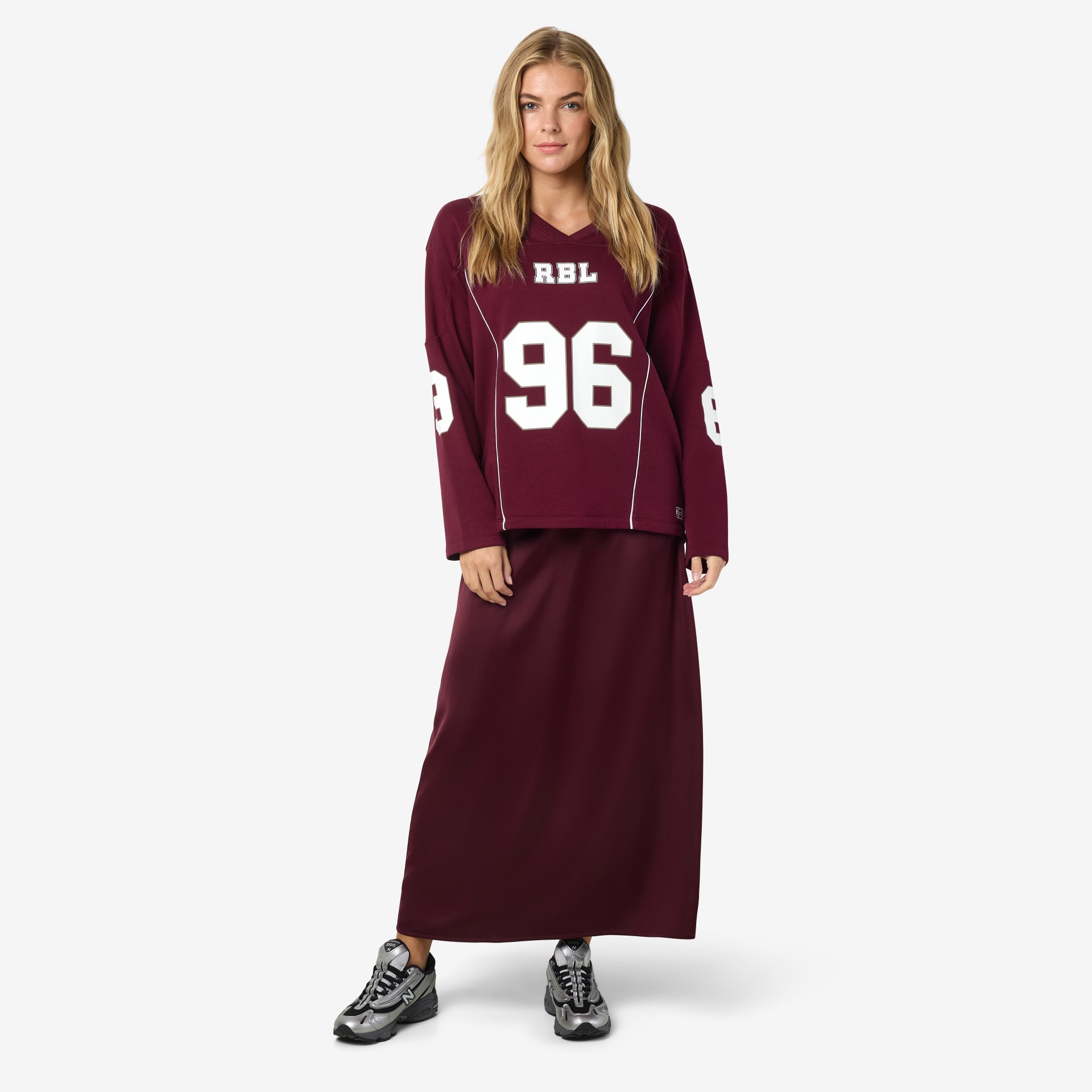 Mulher com sweatshirt vinho com letras RBL e número 96 branco, saia vinho e ténis cinzentos com detalhes pretos e brancos