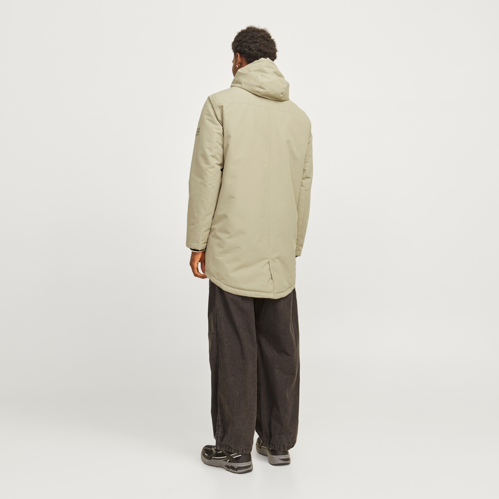 Casaco bege comprido com capuz visto de costas com calças pretas largas e ténis pretos e cinza