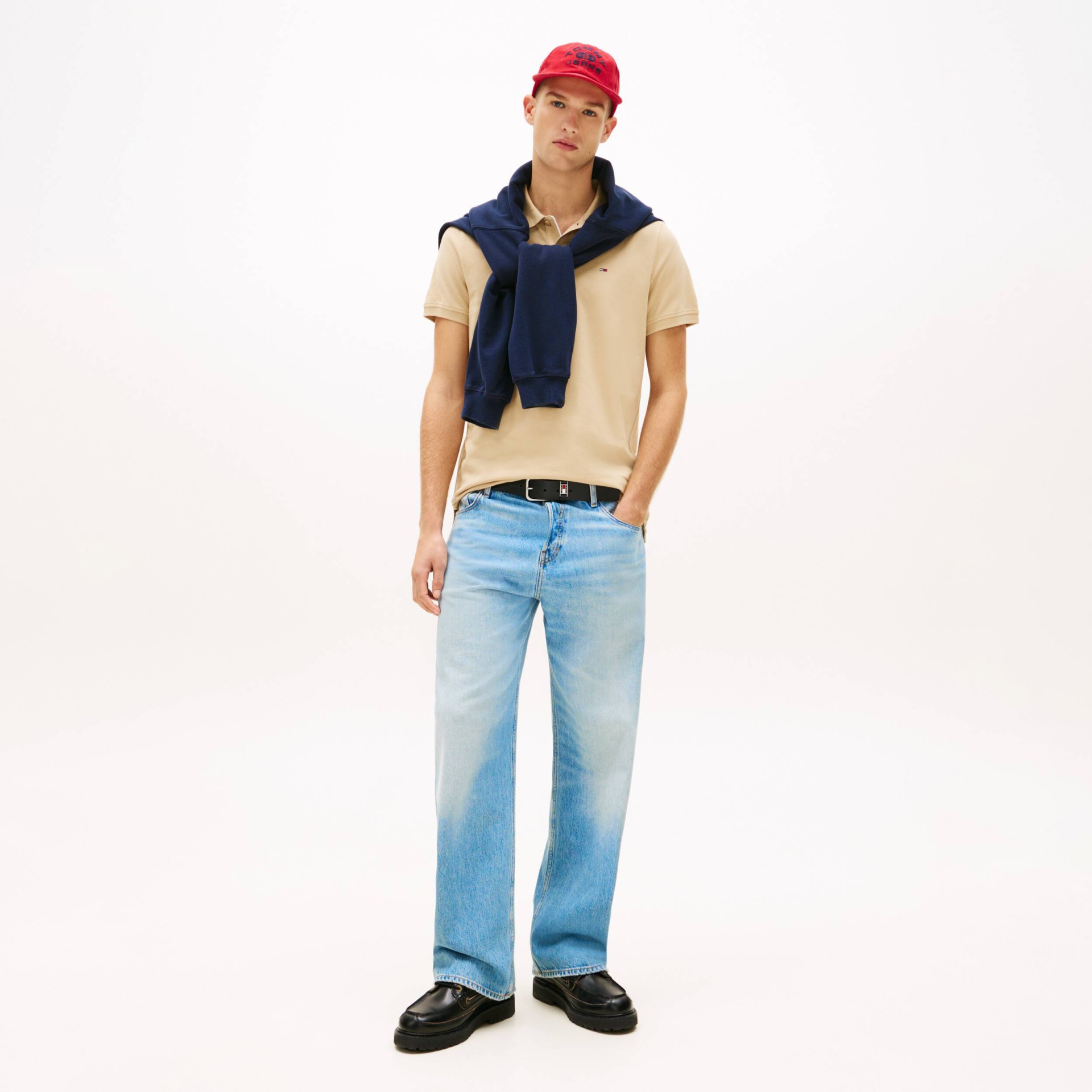 Modelo de homem com roupa casual incluindo boné vermelho, polo bege, camisola azul escura ao ombro, jeans azuis claros e sapatos pretos.